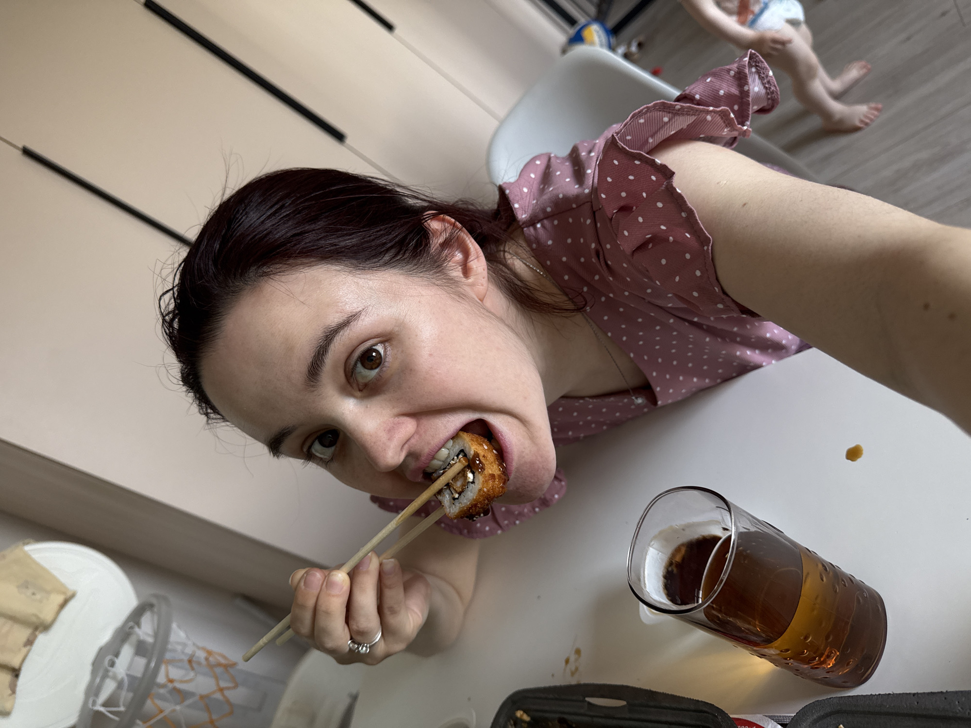 A woman with dark hair pulled back is holding a piece of sushi with chopsticks in her mouth, looking directly at the camera. She is wearing a pink polka-dot shirt. A glass of amber-colored liquid, possibly beer or iced tea, sits on the white table next to her. A sushi container is also on the table. In the background, blurred, are the legs of a small child wearing a diaper. The location is likely indoors, with a white cupboard partially visible on the left and a white chair in the background. It is not possible to determine if this image was taken in Izhevsk, Russia.