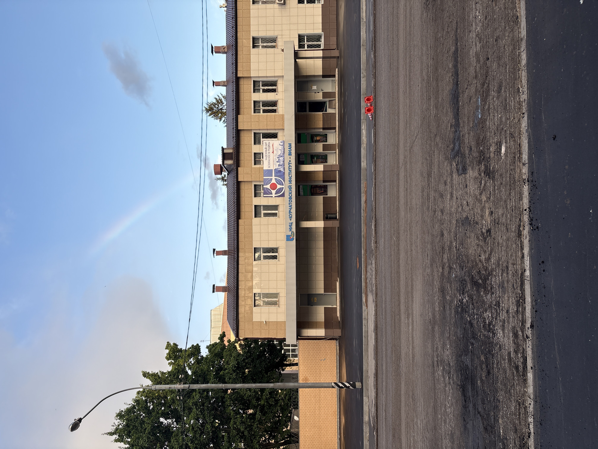 The image captures a frontal view of a two-story building with a light beige and brown facade, located in Moscow, Russia. The building has a dark tiled roof with chimneys. A banner with a blue and red logo and Russian text, "НАДЕЖНЫЙ ЦЕНТР БЕЗОПАСНОСТИ РОДИНЫ", is displayed above the entrance. The building's entrance area features multiple glass doors and windows. In front of the building, a sidewalk is visible, and further in the foreground, a street undergoing apparent repair or construction is shown with loose asphalt and gravel. Two orange traffic cones are placed on the street. To the left of the building, a tall brick wall topped with barbed wire runs along the property line. A mature tree with dense green foliage stands to the left of the wall. In the sky above, a faint rainbow arcs across a partly cloudy blue sky. A lamppost with a curved arm and light fixture is positioned to the left of the building and tree. The overall scene suggests an outdoor view in a city setting.