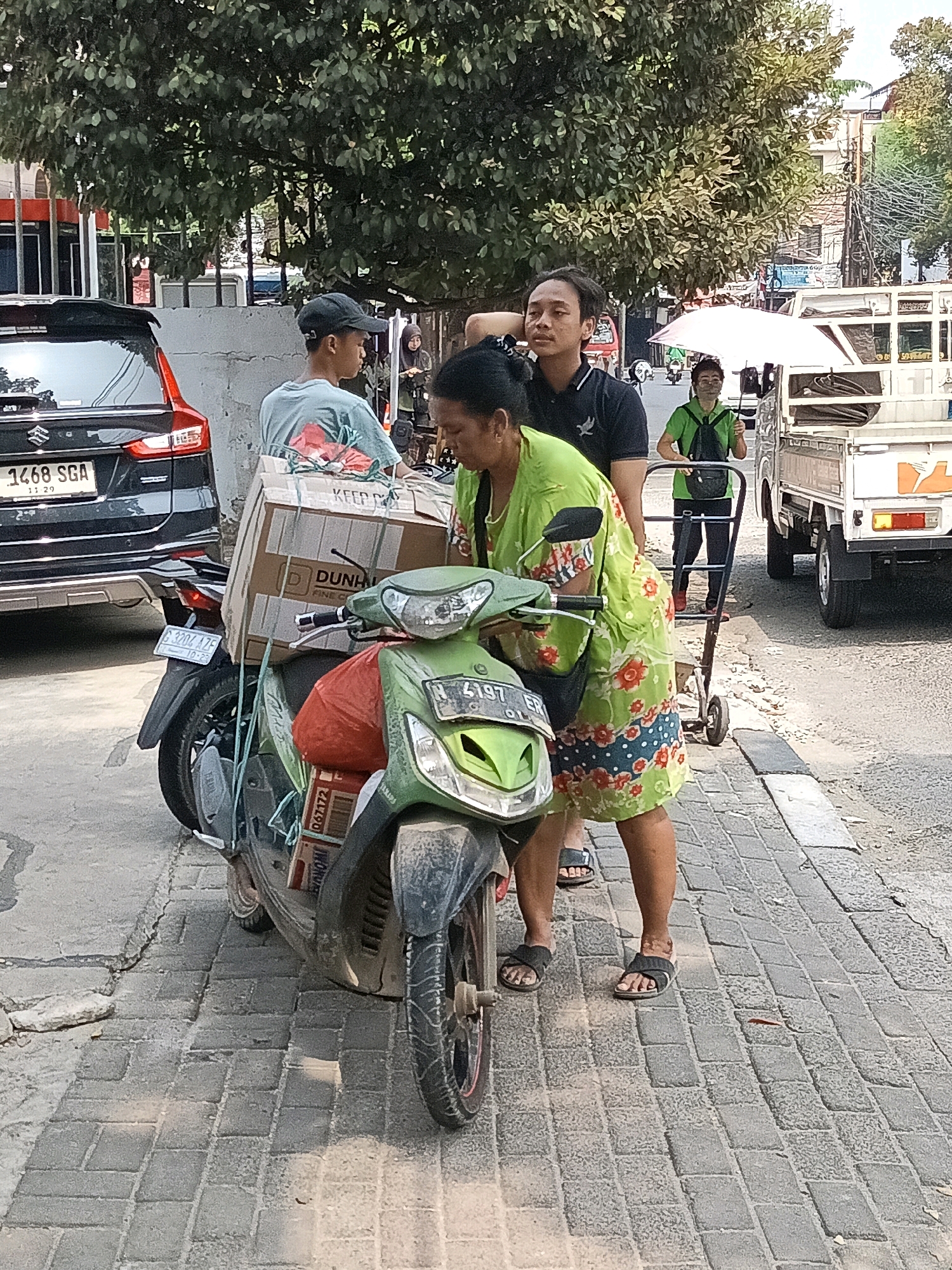 Two individuals are on a motorcycle with a large cardboard box strapped to the back. The motorcycle is green and appears to be a scooter. The cardboard box is marked with "DUNHILL" and "KEEP CLEAN." A woman in a green floral dress and a man in a black shirt are standing next to the motorcycle. A second man, wearing a light blue t-shirt and a black baseball cap, stands behind the car. The scene appears to be a street in South Jakarta, Indonesia, with trees, buildings, and other vehicles visible in the background. The ground is paved with cobblestones.