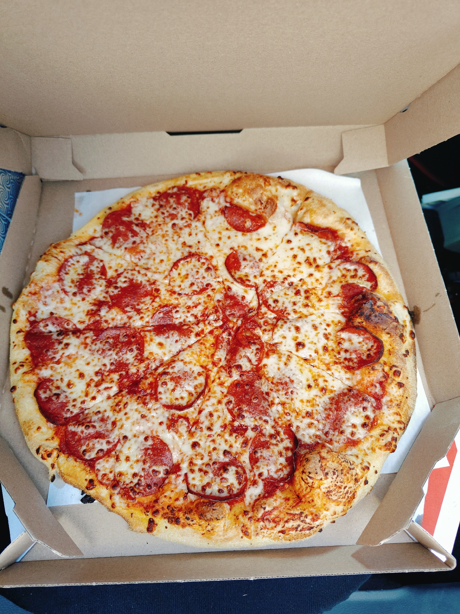 A close-up, overhead shot shows a pepperoni pizza inside an open cardboard pizza box. The pizza is round and appears to be a standard medium size, with a golden-brown crust. It is topped with melted mozzarella cheese and numerous circular slices of pepperoni. The pizza is centered within the box, which is made of plain brown cardboard. The box is resting on a dark surface, possibly a car seat or a table. The background is slightly out of focus, but hints of blue and white patterned material are visible on the left side of the box, and a red and white striped object is partially visible on the right. No people are visible in the image. The lighting suggests it is daytime, and the image appears to be taken indoors or in a shaded outdoor area. The location is Nowy Dwór Mazowiecki, Poland.