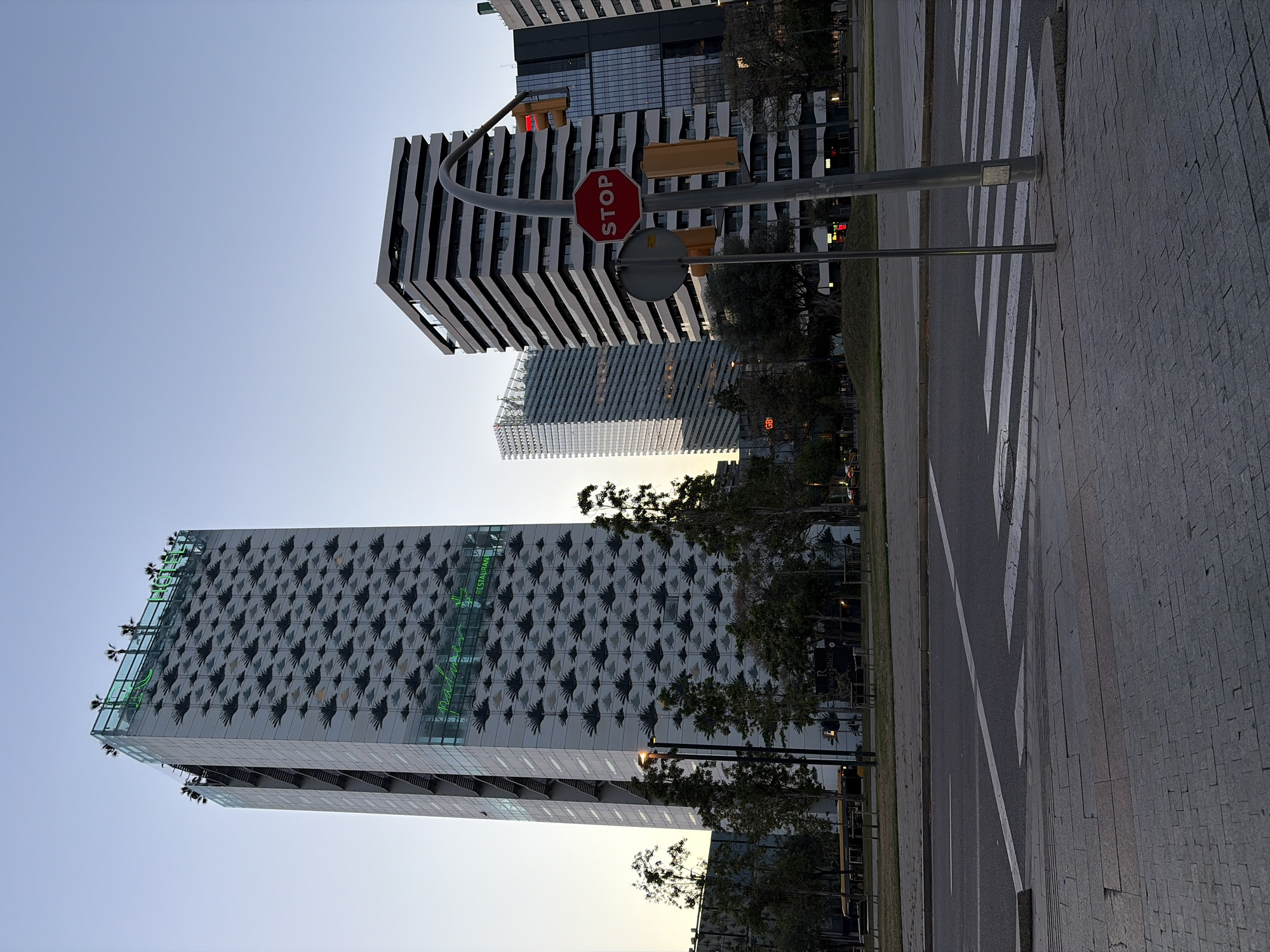 The image displays a modern urban street scene at what appears to be dusk or late afternoon, characterized by clear skies. The photograph is taken from a street-level perspective, looking towards a cluster of contemporary high-rise buildings.
In the foreground, a multi-lane street is visible, featuring a prominent zebra crossing with white stripes. The street is bordered by a paved sidewalk made of dark grey interlocking pavers on the right and a grassy median strip with small trees on the left. A tall grey pole stands on the right side of the street, supporting a red octagonal STOP sign with white lettering. Above the stop sign, a set of yellow-cased traffic lights hangs over the road; one red light is illuminated. Another circular regulatory sign is affixed to the pole, its back facing the viewer.
Several modern buildings dominate the background. On the far left, a tall, light-colored skyscraper features a distinctive facade patterned with numerous dark, stylized shapes resembling birds or leaves. Its upper section includes a rooftop terrace with small trees or plants. Green neon text is visible on this building, spelling "RESTAURANT" horizontally and possibly "mademoiselle" or a similar word vertically. Below the green text, another sign spells "RENAISSANCE".
Towards the center-right, another building showcases a facade of alternating horizontal light and dark bands. Behind it, a third building with a grid-like or geometric pattern is partially visible. Further to the right, a building appears to have a "BBVA" logo, indicating a Spanish bank. The overall setting suggests a commercial or business district in a developed city, consistent with the l'Hospitalet de Llobregat, Spain context. No people or vehicles are explicitly visible, rendering the scene quiet and still.