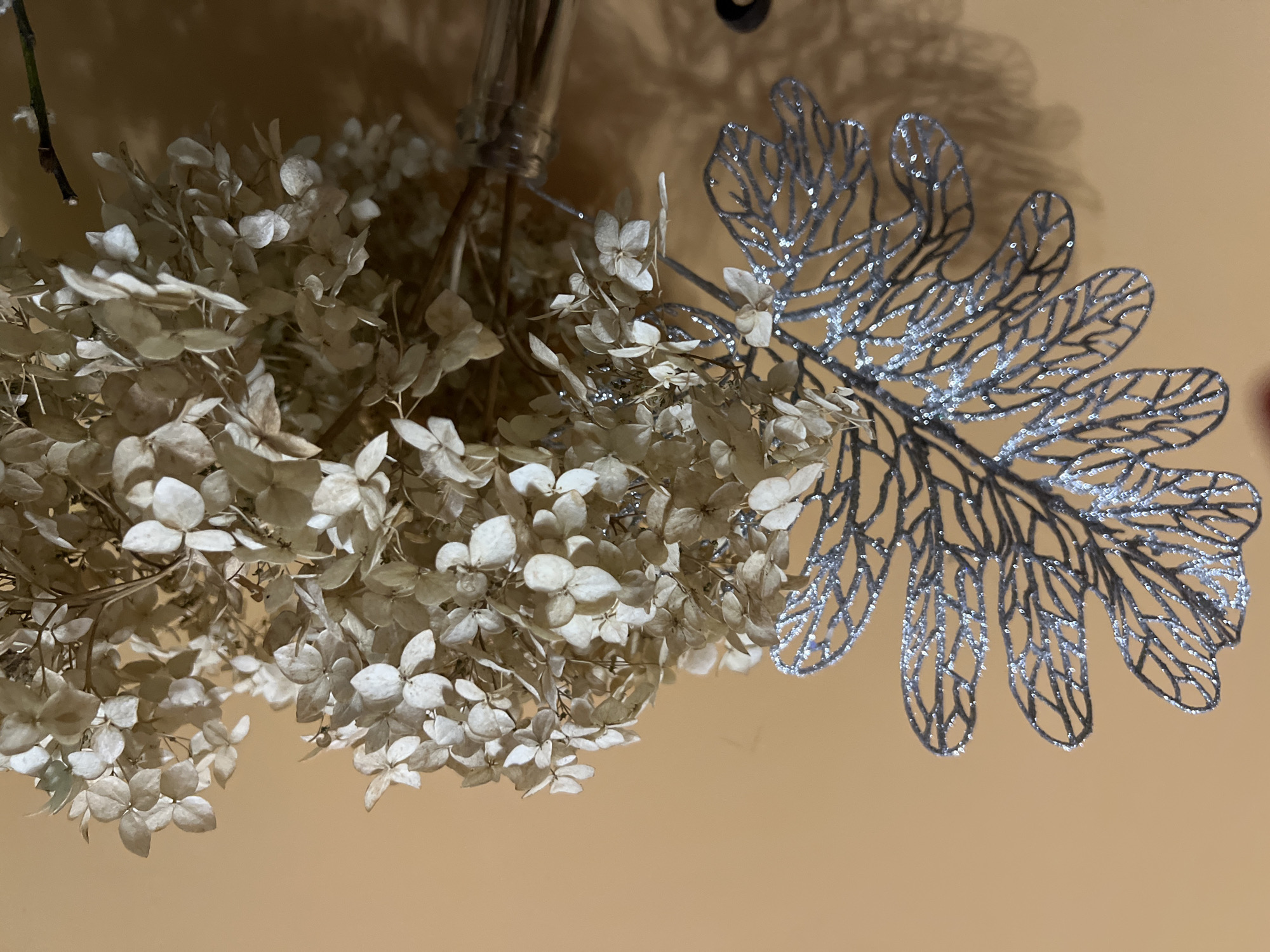 A close-up shot focuses on a metallic silver leaf-shaped ornament with glitter accents, positioned to the left of a bunch of dried hydrangeas. The hydrangeas, in various shades of beige and white, fill the right side of the frame. The arrangement is placed in a clear glass bottle, partially visible at the bottom right. The background is a solid, warm orange wall, with shadows cast from the objects onto it. No people are present in the scene. The image does not provide any specific visual cues to indicate a location within the city of Minsk, Belarus.