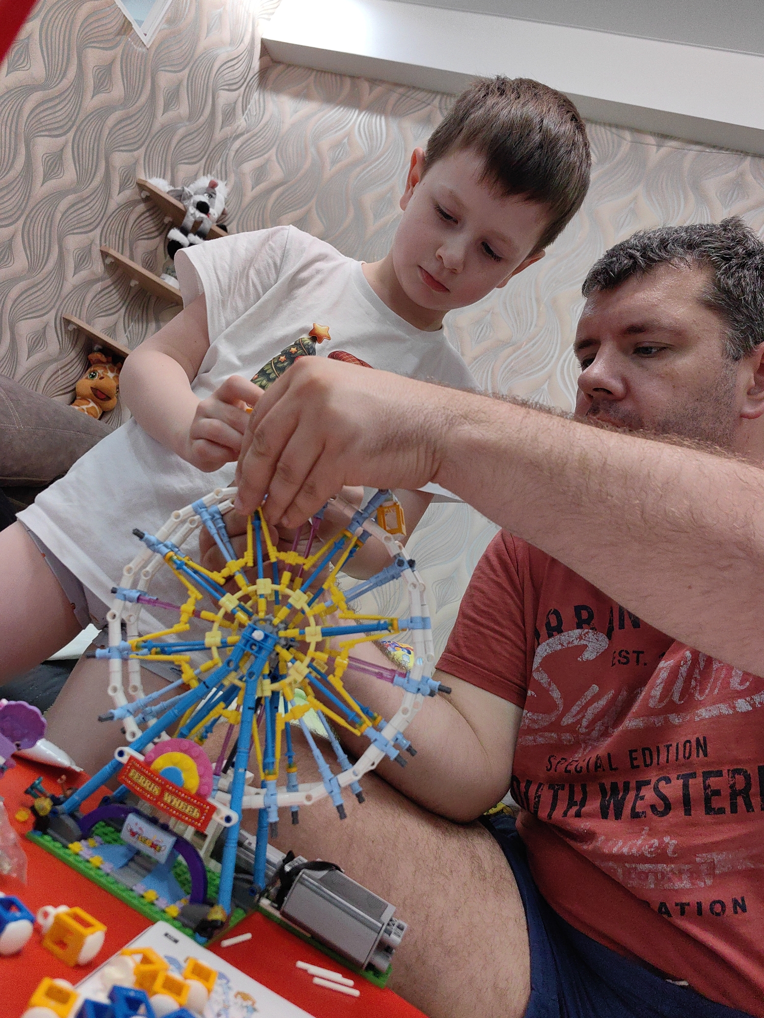 In an indoor environment with textured wallpaper, a man and a young boy are collaboratively assembling a Ferris wheel toy. The man, wearing a red t-shirt with text that includes "Special Edition" and "Western", is focusing on the construction. The boy, in a white t-shirt, is also actively participating in building the toy. Various toy components, including colorful plastic pieces and a phone, are scattered on a red surface in front of them. The setting appears to be a domestic space. There is no identifiable information in the image that suggests the location is the city of Sochi, Russia.