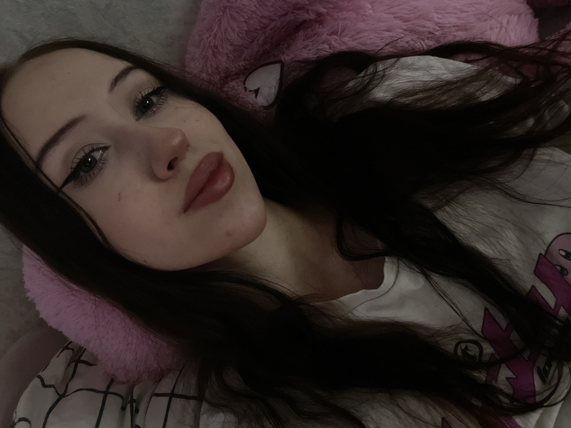 The image shows a close-up, dimly lit selfie of a young woman lying down. She has long, dark hair and is wearing makeup, including eyeliner and lip color. She is wearing a white t-shirt with a graphic print on it. She is lying on what appears to be a bed with pink pillows and bedding. There is no indication of her location in Votkinsk, Russia.