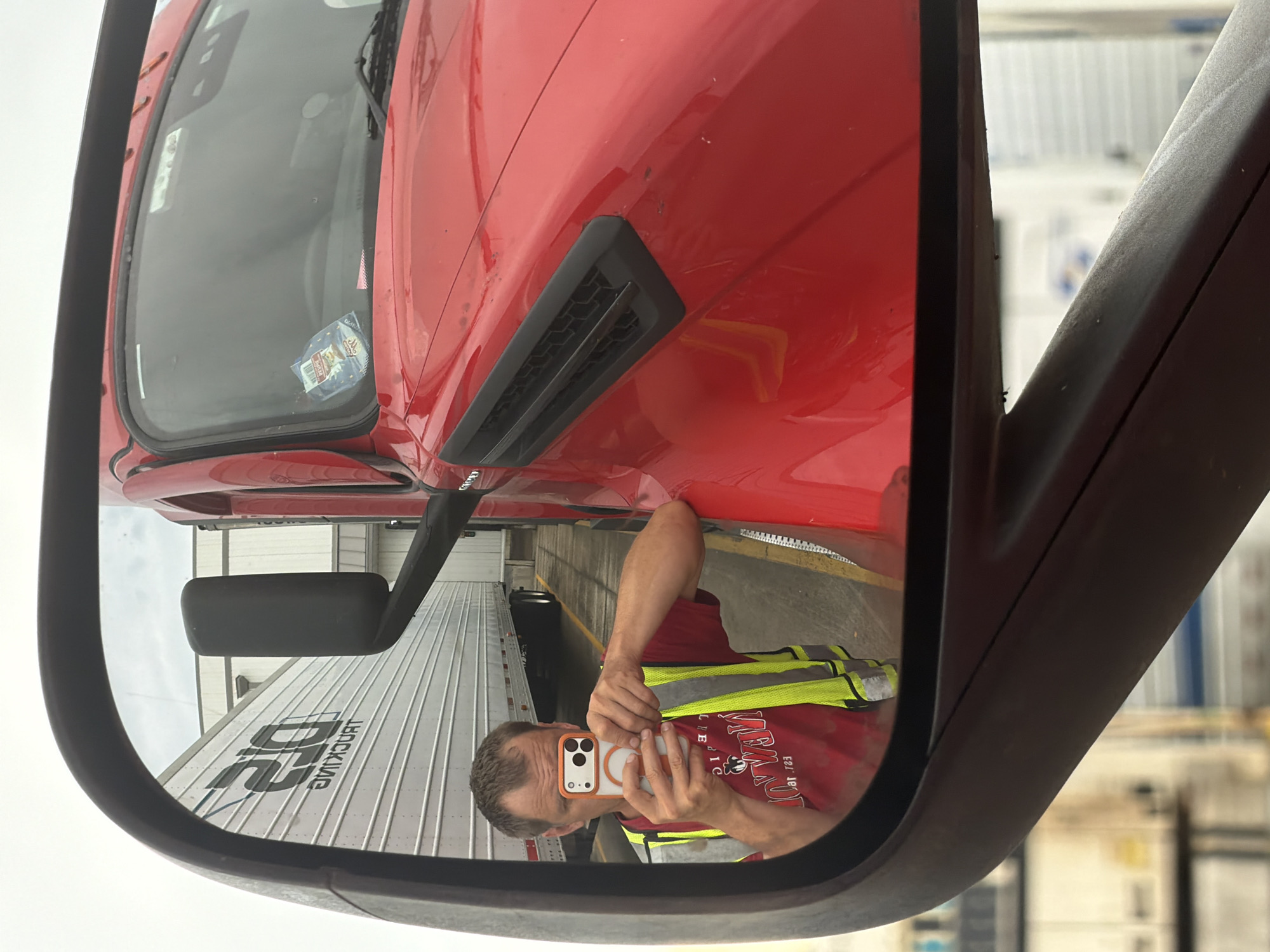 A person is taking a selfie in the side mirror of a red semi-truck. The person is wearing a red shirt and a high-visibility yellow vest. They are holding an orange-cased smartphone in their right hand. In the reflection of the mirror, other semi-trucks and trailers are visible, along with a building structure in the background. The ground appears to be wet, suggesting recent rain. The scene takes place outdoors in what appears to be a truck yard or industrial area. The image has a slightly overcast sky. The location is not explicitly stated in the image.