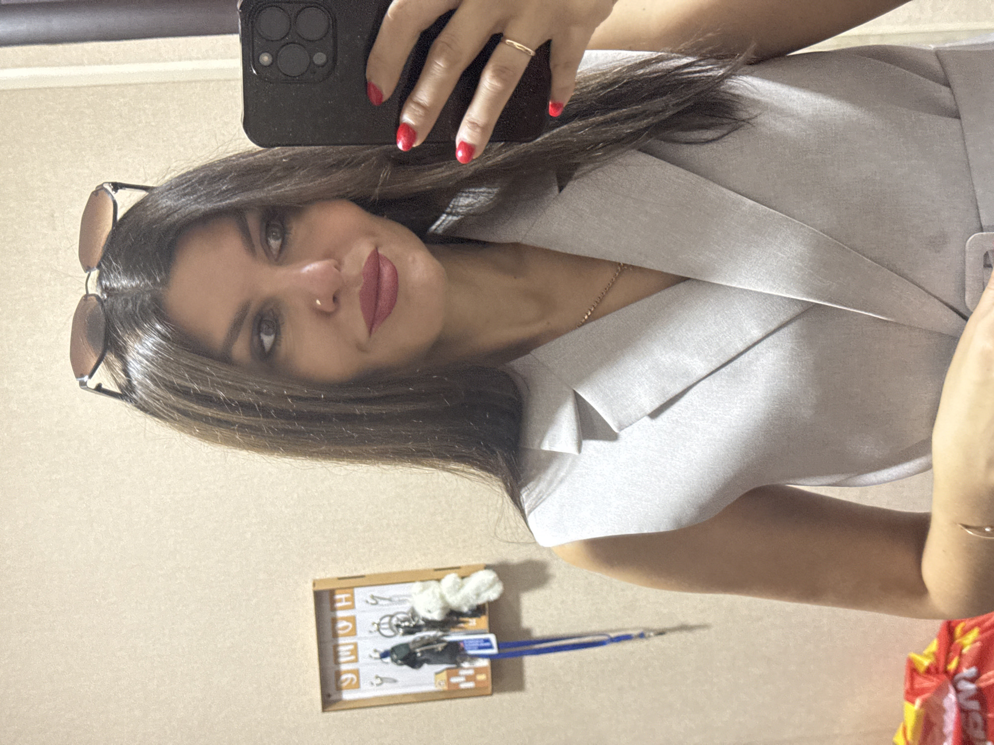A woman is taking a mirror selfie in an indoor setting. She has long, dark brown hair with sunglasses perched on top of her head. She is wearing a light gray sleeveless blazer and has red nail polish on her fingernails. She is also wearing a delicate gold chain necklace and a thin gold ring on her left hand. To her left, a wooden key holder with the word "HOME" in wooden letters is mounted on a light-colored wall. Several keys and two small keychains, one a white teddy bear, hang from the hooks. The overall environment appears to be a residential interior, possibly an entryway or hallway. The image does not contain information that specifies the location within the city of Chaikovsky, Russia.