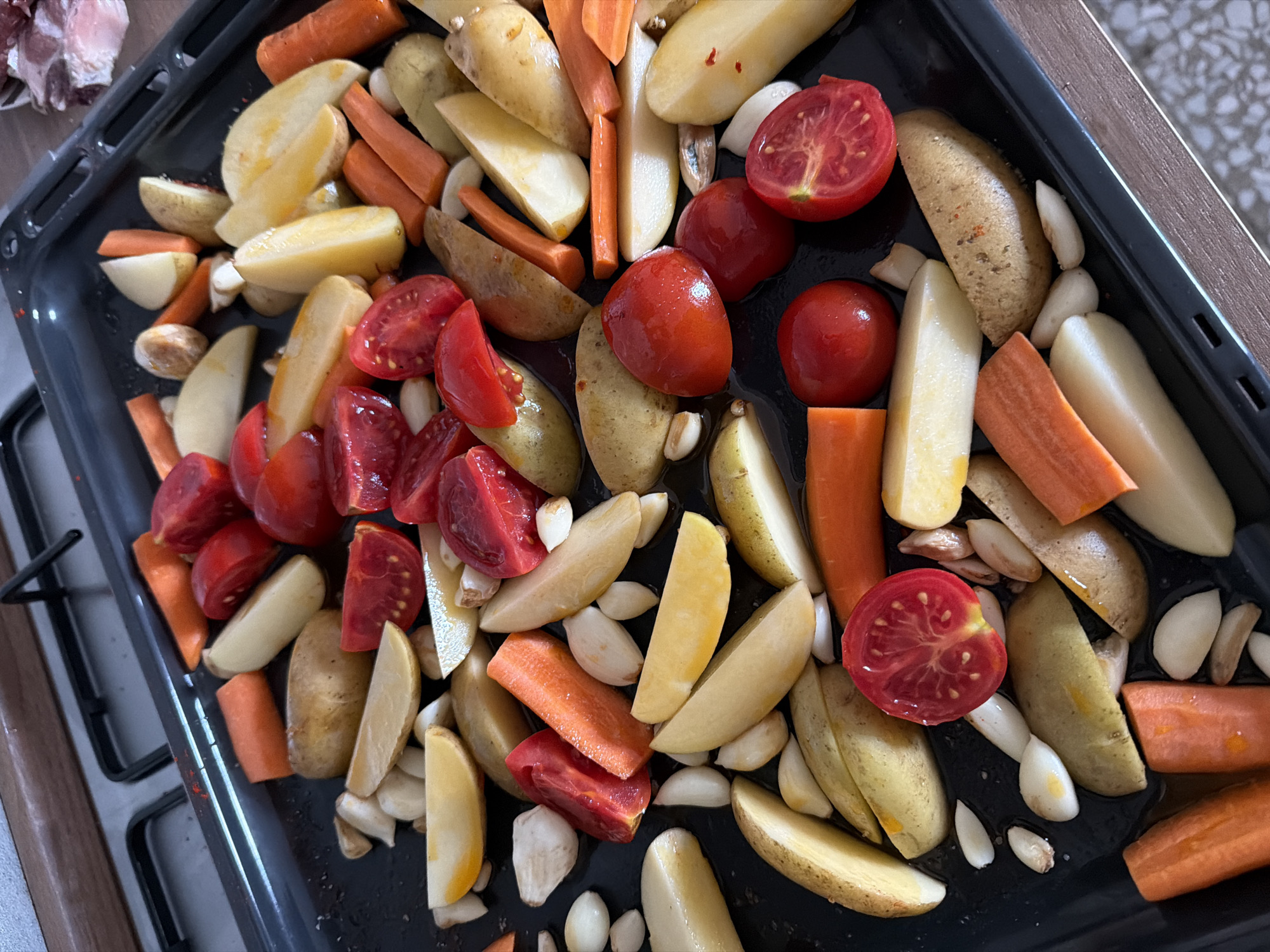A close-up shot reveals a black baking tray filled with an assortment of raw vegetables, likely prepped for roasting. The vegetables include: halved red tomatoes, peeled and sliced carrots, wedges of potatoes with skin on, and scattered cloves of garlic. The tray appears to have a slight sheen, possibly from oil or other liquid. The tray sits on what appears to be a dark metal rack, which is likely part of a stove or oven. The edge of a countertop of some sort is partially visible to the right of the tray. The location cannot be determined from this image alone.