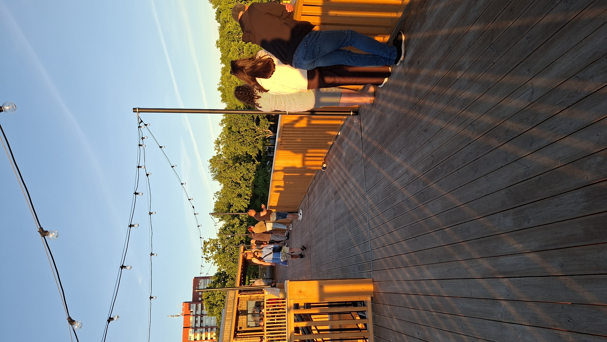 The image depicts a wooden outdoor deck, likely a restaurant or bar, in Tallinn, Estonia. The scene is bathed in the warm glow of the setting sun, casting long shadows across the weathered wooden planks. In the background, lush green trees form a dense line, with a red apartment building visible further in the distance. A string of decorative lights stretches across the top of the frame, adding to the ambiance.
Several people are scattered across the deck, appearing relaxed and engaged in conversation or simply enjoying the view. One person walks away from the camera, while others stand in small groups, leaning on the wooden railing that borders the deck. A few individuals are looking out towards the trees, possibly admiring the scenery. The overall impression is one of a pleasant evening gathering in a scenic urban setting.