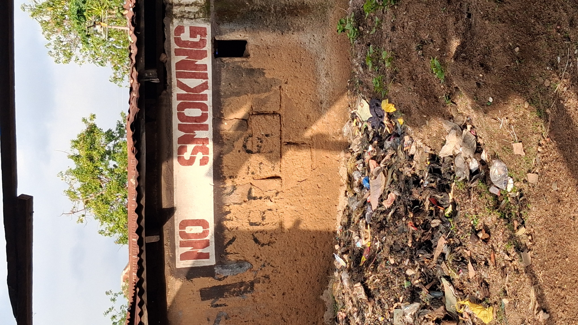 The image depicts a weathered, mud-walled structure with a corrugated metal roof. A white sign with red stenciled lettering reads "NO SMOKING". The wall below the sign is rough and shows signs of age, with some darker smudges that could be old paint or water damage. There is a small, dark rectangular opening in the wall to the right of the sign, resembling a vent or small window. In front of the structure, a large pile of refuse is visible, consisting of various types of trash including plastic bags, papers, and what appears to be organic waste, indicating a general area of disrepair or a refuse collection point. The ground in the foreground is a mixture of dirt and scattered debris. The background shows green trees and a cloudy sky. This scene is located in Jos, Nigeria, suggested by the overall appearance and context of the image.