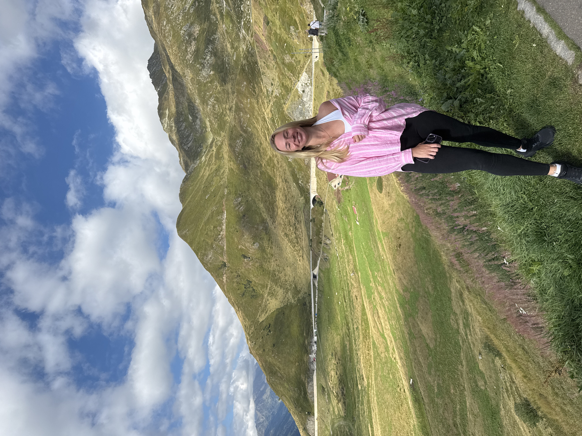 A young Caucasian woman with blonde hair, wearing a pink and white striped unbuttoned shirt over a white sports bra and black leggings, stands facing the camera on a grassy slope. She holds a black smartphone in her right hand. Her left arm is relaxed by her side. Behind her, a lush green mountain landscape unfolds under a partly cloudy blue sky. A road snakes through the valley, with a tunnel visible. In the distance, other mountain ranges are visible. Two figures are seen walking on the road in the background. The woman is smiling. This scene is located in Airolo, Switzerland.