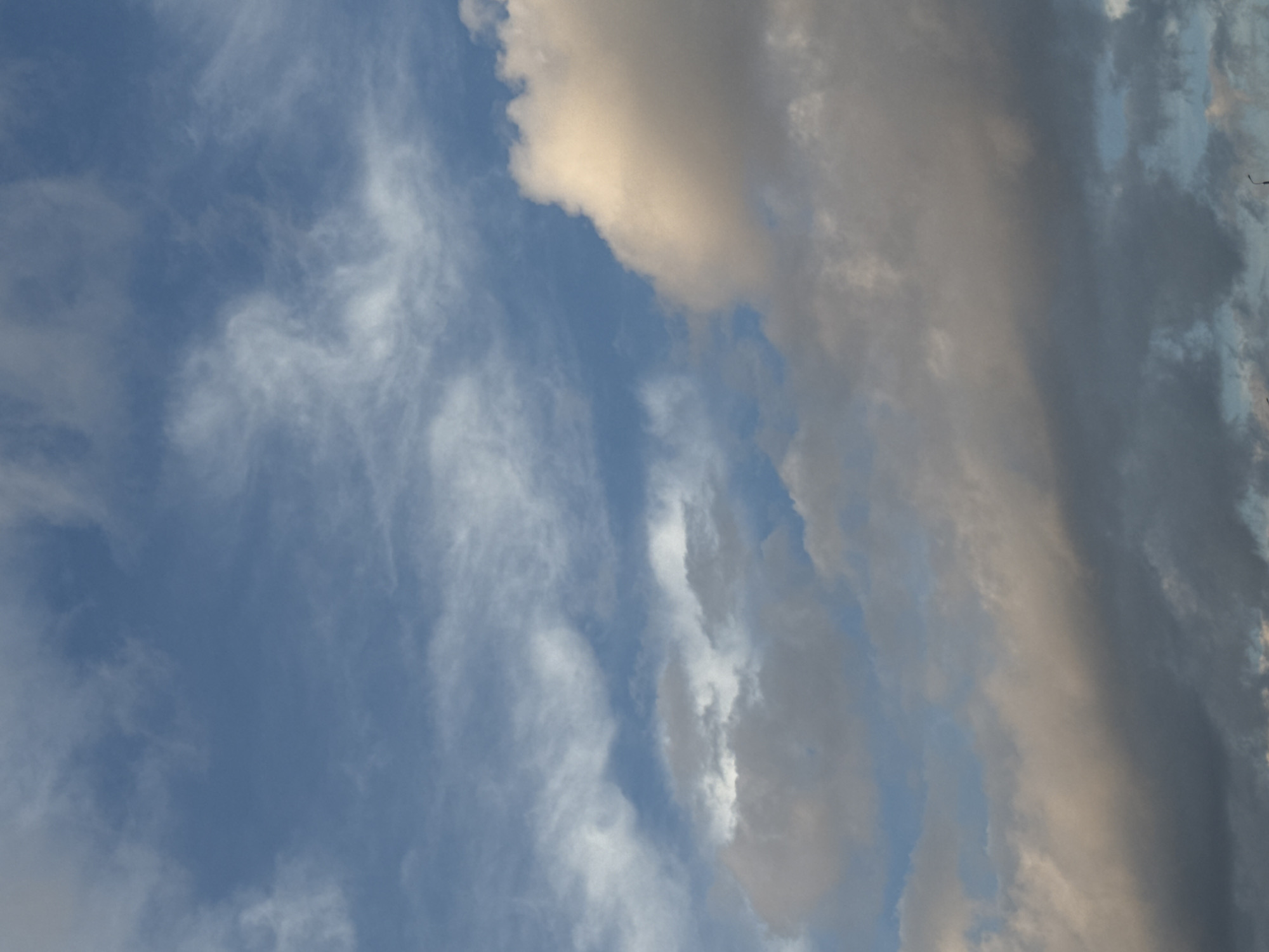 The image shows a sky filled with clouds. The left side of the image features wispy, white cirrus clouds against a blue sky. On the right, there are darker, more layered cumulonimbus clouds in shades of grey and beige. The location is not evident from the image.