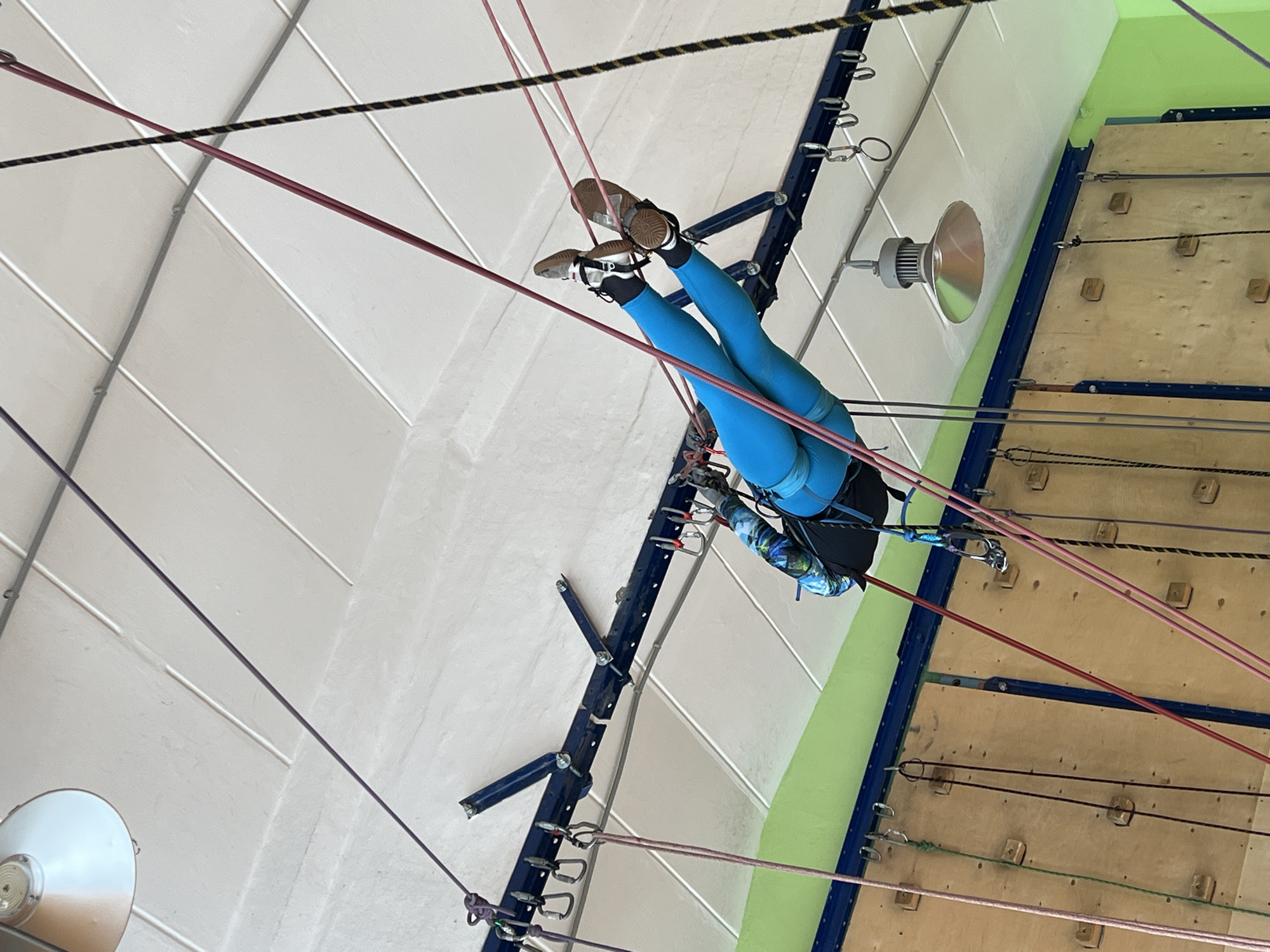 A person is suspended upside down by ropes in what appears to be an indoor climbing facility. The individual is wearing blue leggings, shoes, and a harness with attached climbing gear. Several ropes of varying colors (red, purple, black and yellow) crisscross the frame, connected to the ceiling and the climbing wall.
The climbing wall, constructed of wood panels, is visible to the right of the person. Blue metal supports frame the wall, and climbing holds are sparsely placed across the surface. A green stripe is painted along the wall's edge.
The ceiling is white with a grid-like pattern of recessed lights. Metal support beams with clips are visible along the edges of the ceiling.
The location is likely in Chaikovsky, Russia, based on contextual information.