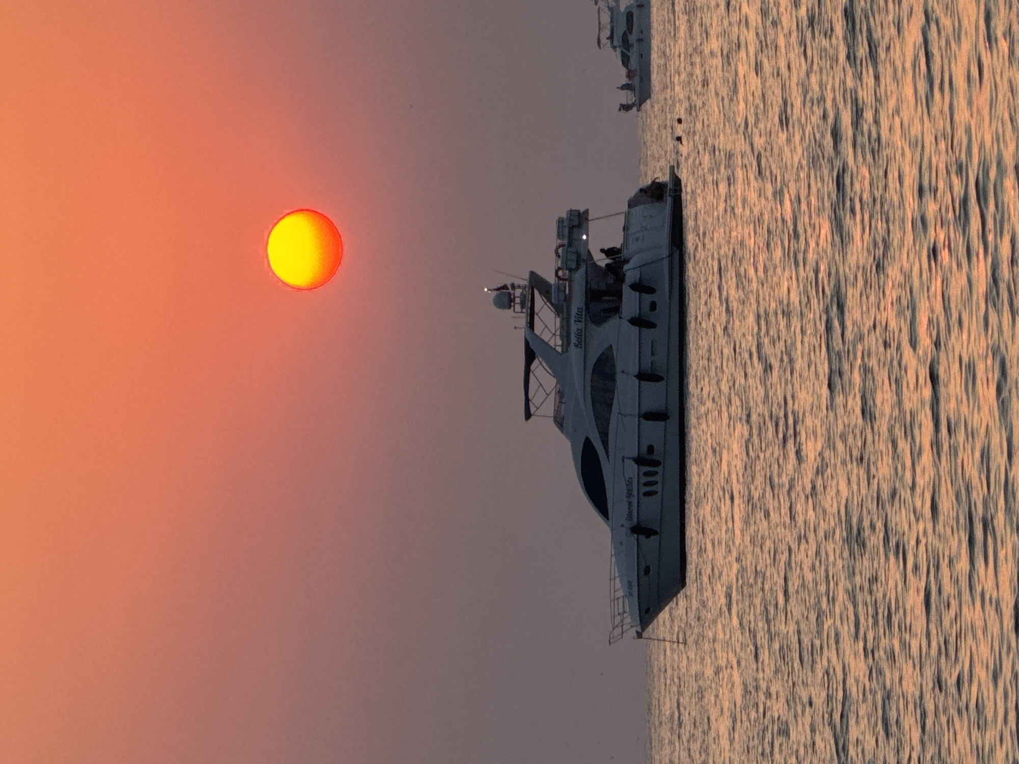 The image depicts a serene sunset scene over the waters of Dubai, United Arab Emirates. A large, white yacht, identified by the name "Bella Vita" on its side, is prominently featured in the foreground, anchored on calm, rippling water that reflects the warm hues of the setting sun. Several people are visible on the deck of the yacht, appearing relaxed and enjoying the view. In the distance, another smaller yacht is also present, with a few individuals on its deck. The sky is a gradient of vibrant orange and pink, with the sun, a bright yellow orb, positioned in the upper right quadrant of the frame. The overall atmosphere is tranquil and picturesque, characteristic of Dubai's coastal beauty during twilight.