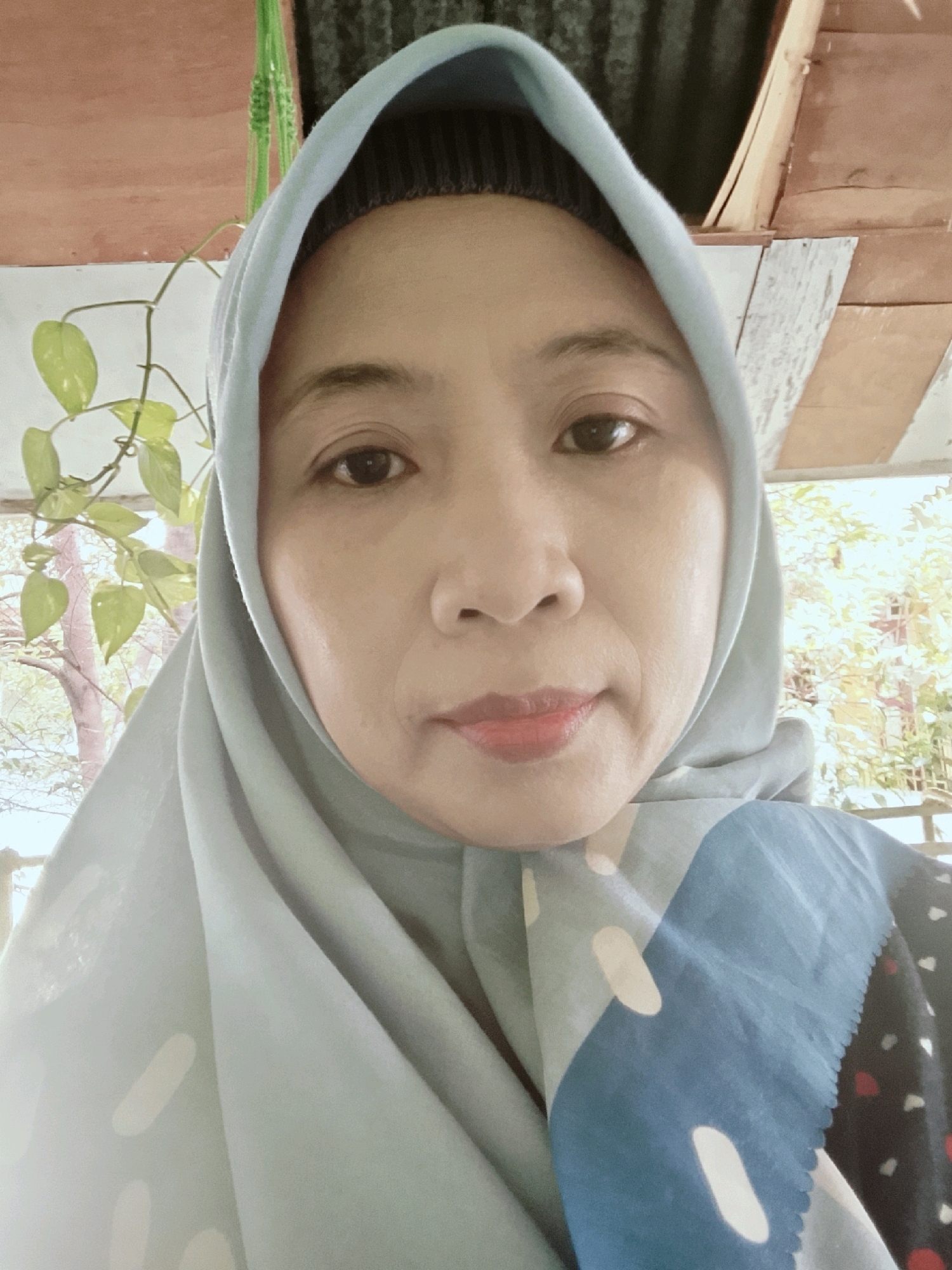 An Indonesian woman with fair skin and dark eyes wears a light blue hijab and a patterned garment. She is positioned in front of a backdrop featuring wooden structures and green foliage, suggesting an outdoor or semi-outdoor setting. The image appears to be a selfie, with the woman's face as the central focus. The environment, with its natural elements and rustic wooden construction, could be characteristic of a residential area in Besusu Timur, Indonesia, where traditional or naturalistic architecture is common.