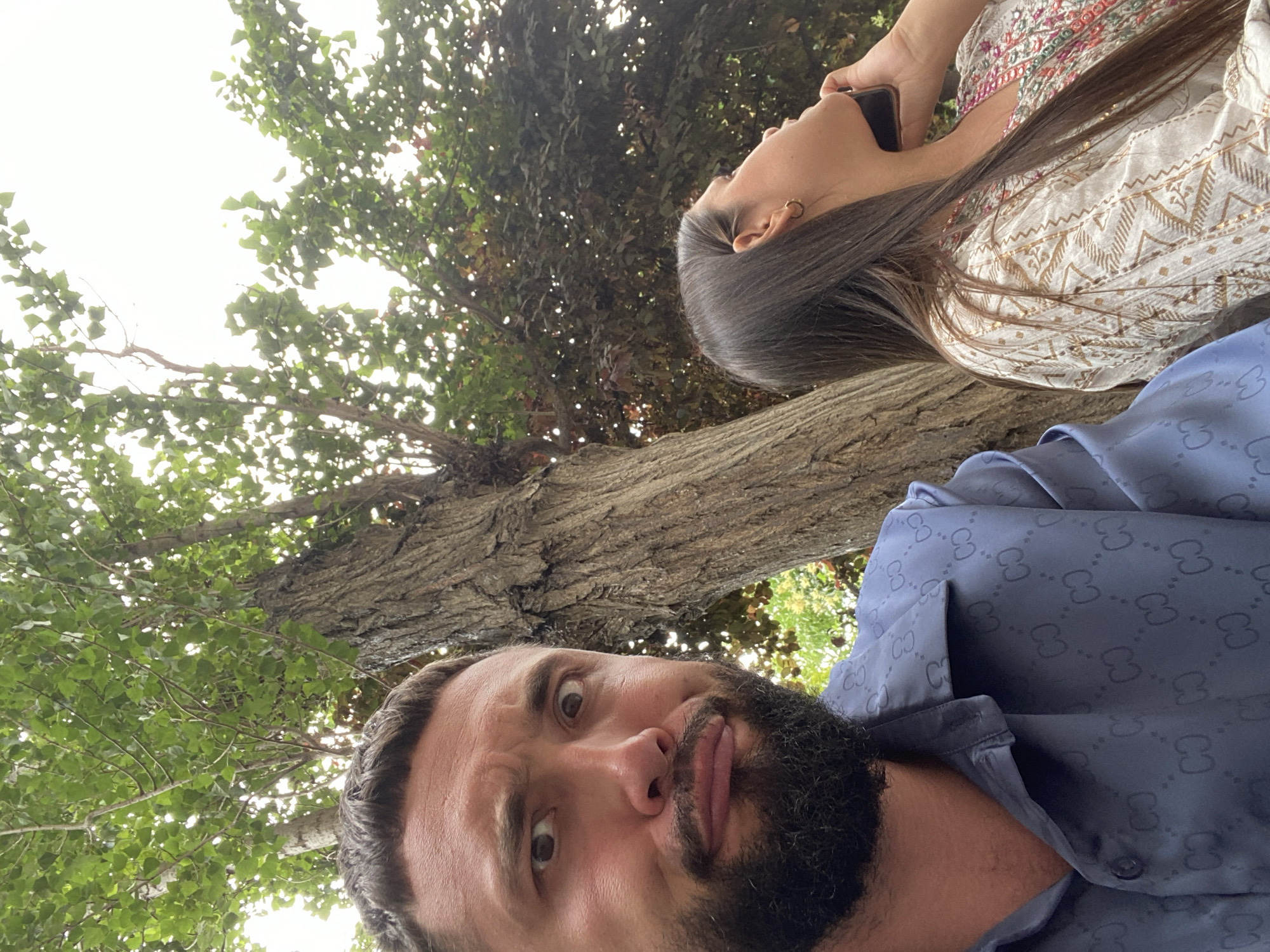 In this selfie-style image taken outdoors, a man with a beard makes a humorous or surprised facial expression towards the camera, looking to his left. He is wearing a blue patterned shirt. To his right, a woman is partially visible, looking upwards and talking on a mobile phone, holding it to her ear. She is wearing a white shirt with colorful embroidery. They are both seated outdoors, with the focus on their faces and upper bodies. Behind them, a large tree trunk is prominent, with green and darker foliage forming the background, suggesting a park or garden setting. The overall lighting is bright, with the sky visible through the leaves. The location appears to be a public outdoor space within the city of Derbent, Russia, likely a park or a seating area near a natural landscape.