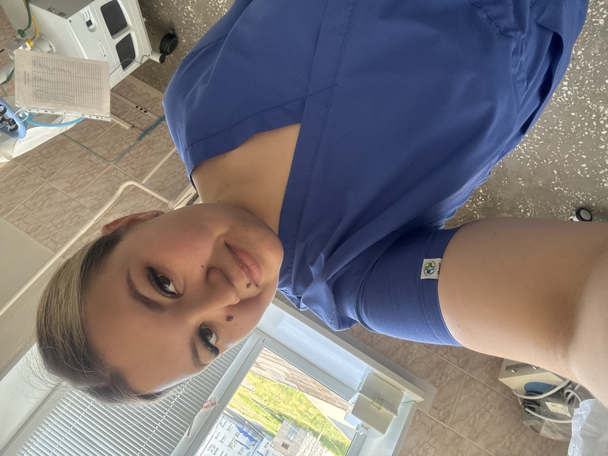 A selfie taken by a woman wearing blue scrubs inside what appears to be a medical facility. She has blonde hair pulled back and is smiling at the camera. There are medical devices and equipment visible in the background, including a ventilator and various monitors. A window shows a view of buildings and greenery, suggesting an urban or suburban setting. The specific location in Chaikovsky, Russia, cannot be determined from the image alone.