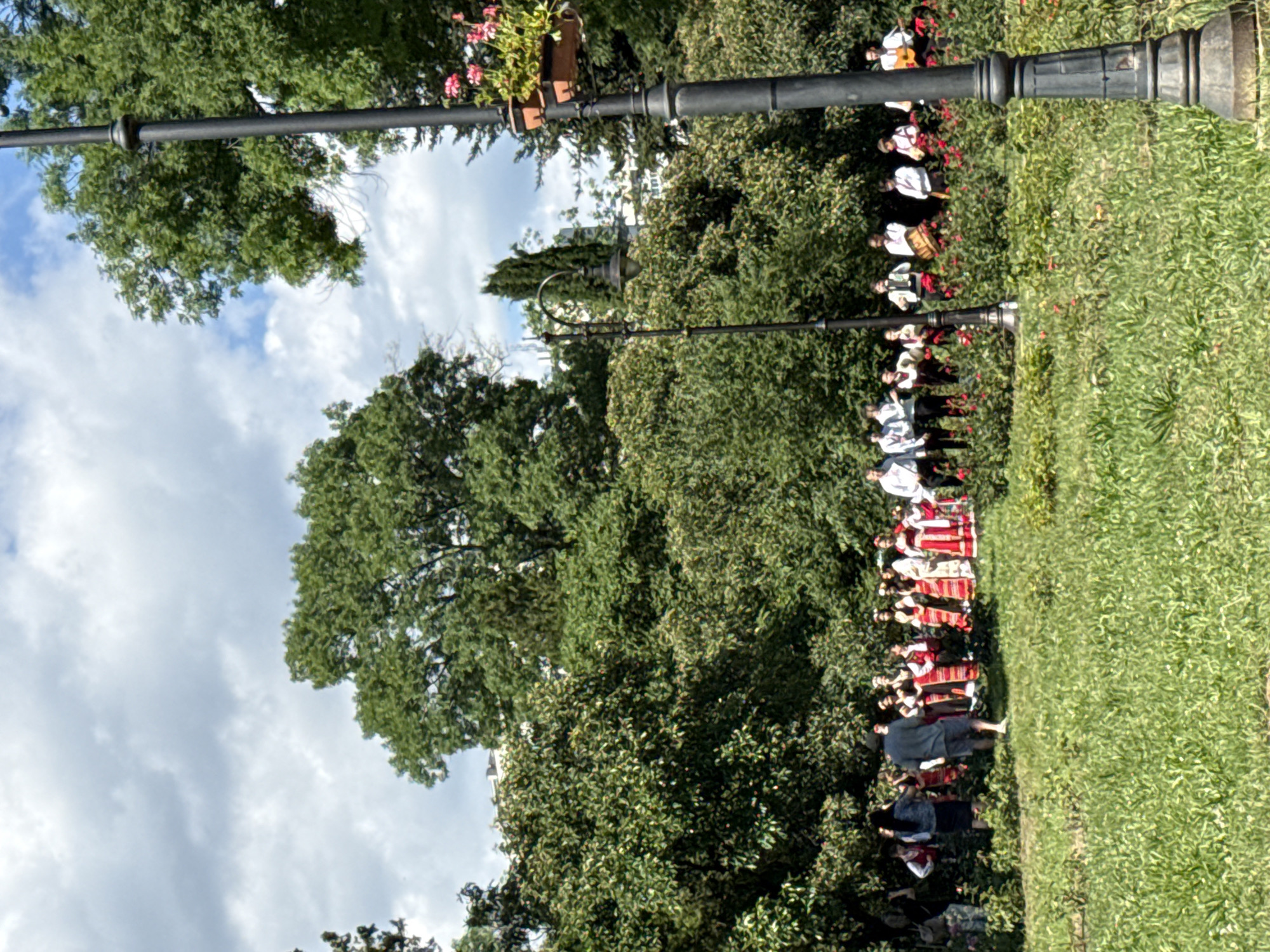 In an outdoor park setting with lush green trees and grass, a group of people are gathered. Some are dressed in traditional folk costumes, including long skirts with patterned designs and embroidered tops. Several individuals are holding musical instruments such as drums and guitars. A few people are observing the group. A black lamppost with a decorative top and a planter with flowers on it is positioned in the foreground on the right side of the image. Another lamppost with an older design is visible to the left. The sky is partly cloudy. The scene appears to be a performance or cultural gathering in a city park.