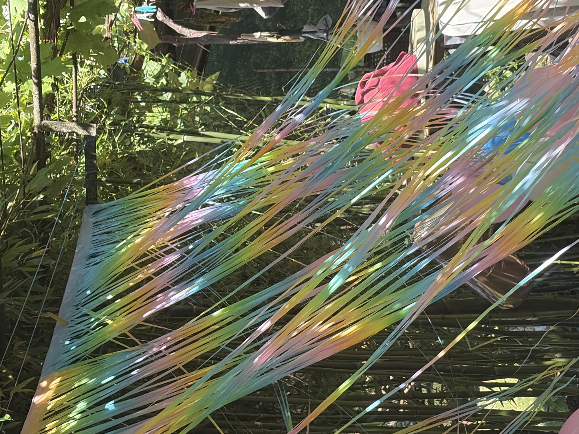 The image shows a close-up of a colorful party decoration made of iridescent streamers hanging outdoors, likely in the city of Vlasatitsa, Bulgaria. The streamers are in shades of pastel pink, yellow, blue, and green, creating a rainbow effect. They are suspended from a wire or string, which is visible at the top of the frame, and blow in the wind, giving a sense of movement. In the background, lush green foliage, possibly bamboo or other dense vegetation, provides a natural backdrop. Hints of a rustic outdoor setting are visible, including a wooden structure or pergola and what appears to be a pink piece of fabric or a large umbrella. The lighting suggests it is daytime with sunlight filtering through the leaves and streamers, creating dappled light and shadows. There are no people visible in the image.