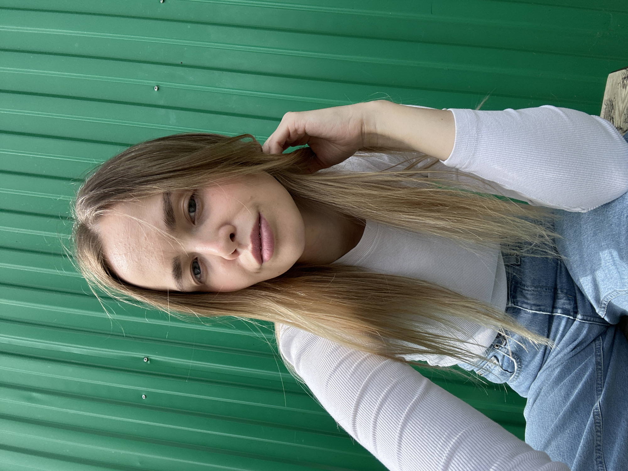 A portrait-style image captures a young woman posing outdoors in Азинский, Russia. The image is rotated 90 degrees clockwise, showing the woman's head tilted towards the left, looking directly at the camera. She has long, straight, light brown or blonde hair, which cascades over her shoulders and chest. Her right hand is raised, with fingers gently touching her hair near her temple.
She is wearing a white, long-sleeved, ribbed top and blue denim jeans. The ribbed texture of her top is clearly visible, especially on the sleeves. Her expression is neutral with a slight hint of a smile, and she has visible pink lipstick.
The background consists primarily of a bright, vibrant green corrugated metal surface, likely a fence or the side of a building, which features horizontal ridges and a few visible screw heads. On the right side of the frame, a small portion of a light-colored wooden surface, possibly part of a bench or seating, is visible. The lighting is bright and natural, suggesting it is daytime. There is no visible text in the image.