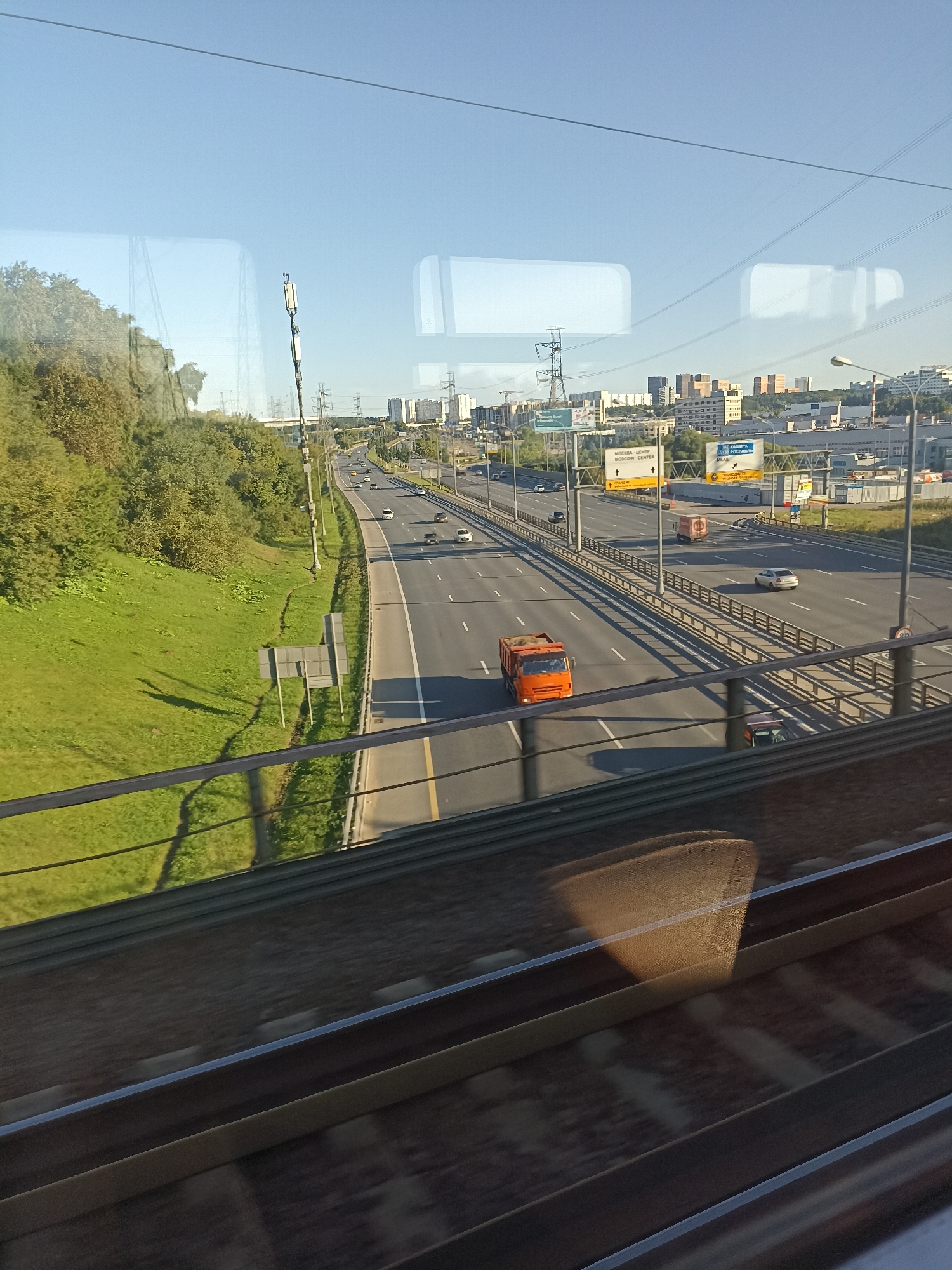 The image shows a view from a train window of a multi-lane highway in Moscow, Russia. The highway is bordered by a grassy embankment on the left and by barriers and other vehicles on the right. Several cars and trucks are visible on the highway, moving in both directions. In the background, there are buildings and infrastructure, including power lines and cell towers. The sky is clear and blue. The foreground of the image shows the interior of the train, including reflections on the window. The overall scene depicts a typical urban highway environment in Moscow.