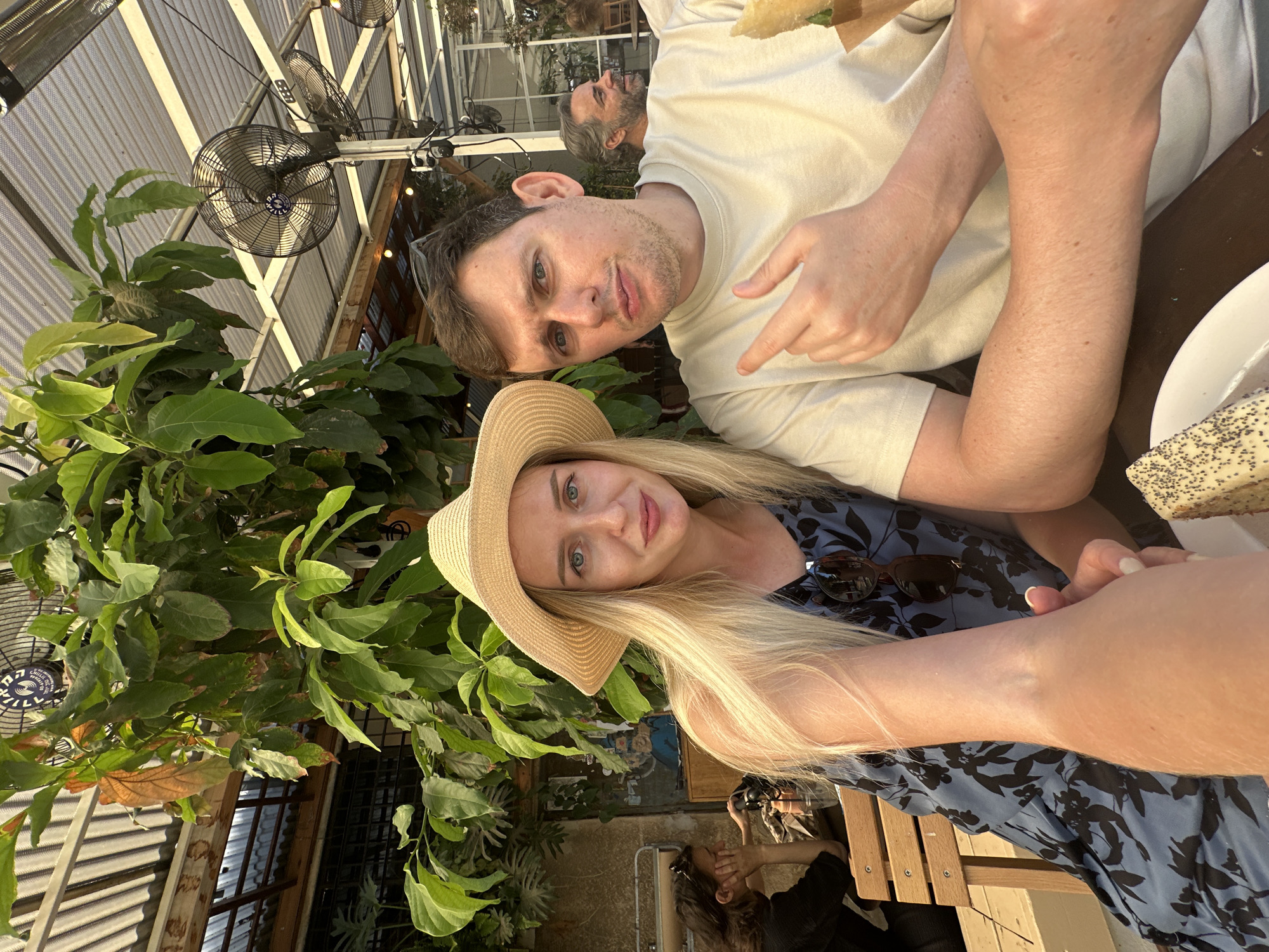 A young couple, a blonde woman wearing a straw hat and a blue floral dress, and a man with short brown hair, are seated at a table outdoors, possibly at a cafe in Tel Aviv, Israel. The woman is on the left, her arm extended towards the camera, while the man is on the right, looking towards the camera with a playful expression and pointing his index finger. Behind them, lush green foliage hangs from above, and industrial fans are visible on the ceiling. Another couple is seated at a table in the background, one of them looking away from the camera. The overall atmosphere suggests a casual, sunny day.