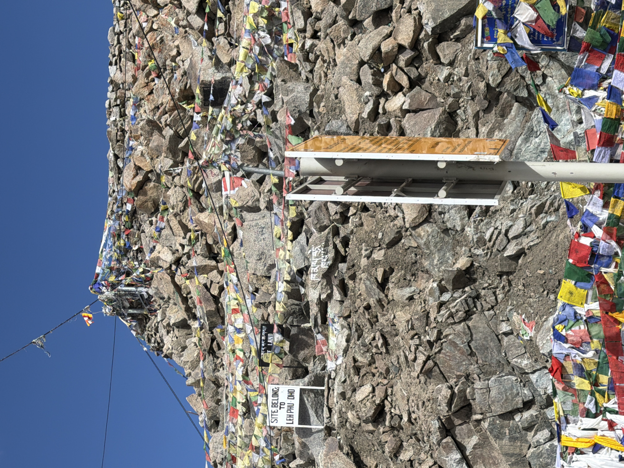 The image shows a mountainous landscape covered with prayer flags. The flags are strung across the rocks and appear to be a significant feature of the location. There are two signs visible. One sign is white with black text that reads "SITE BELONGS TO LEH PHU DHO". Another sign is black with text that appears to be in Tibetan. A vertical metal pole with two signs attached, one white and one orange, is also present in the foreground. The sky is a clear blue. The location is a rocky, elevated area, likely a mountain pass or viewpoint. The presence of prayer flags and signs in Tibetan suggests a connection to Buddhism and the Himalayan region. This scene is consistent with a location in Leh, India, which is known for its Buddhist monasteries and mountainous terrain.