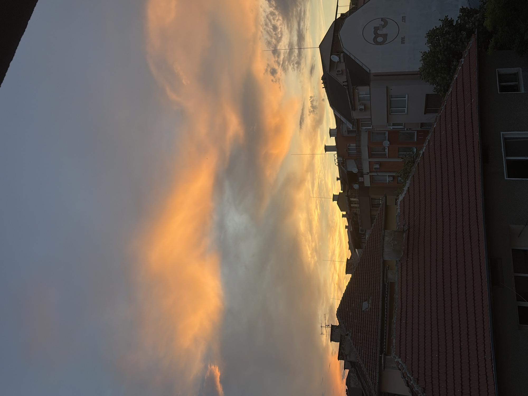 This is an eye-level, medium shot captured from a rooftop vantage point, looking out over a cityscape at sunset. The sky is a dramatic display of color, dominated by vibrant orange and yellow hues from the setting sun, with softer blues and grays in the upper portion. Wispy clouds are scattered throughout, catching the light and adding texture to the sky.
In the foreground, the terracotta-tiled roofs of several buildings are visible. These roofs are aged, with some showing signs of wear and tear. Chimneys and ventilation pipes protrude from the rooftops. Further back, a row of apartment buildings stands, varying in height and architectural style. One building on the right has a prominent white facade with a circular logo and the text "WWW. .COM". Numerous satellite dishes are affixed to the sides of some buildings.
The overall scene depicts a residential urban environment transitioning from day to night, characterized by the warm glow of the sunset illuminating the architecture. There are no people visible in the image, and no explicit actions or interactions are taking place. The environment is quiet and still, emphasizing the natural spectacle of the sunset.