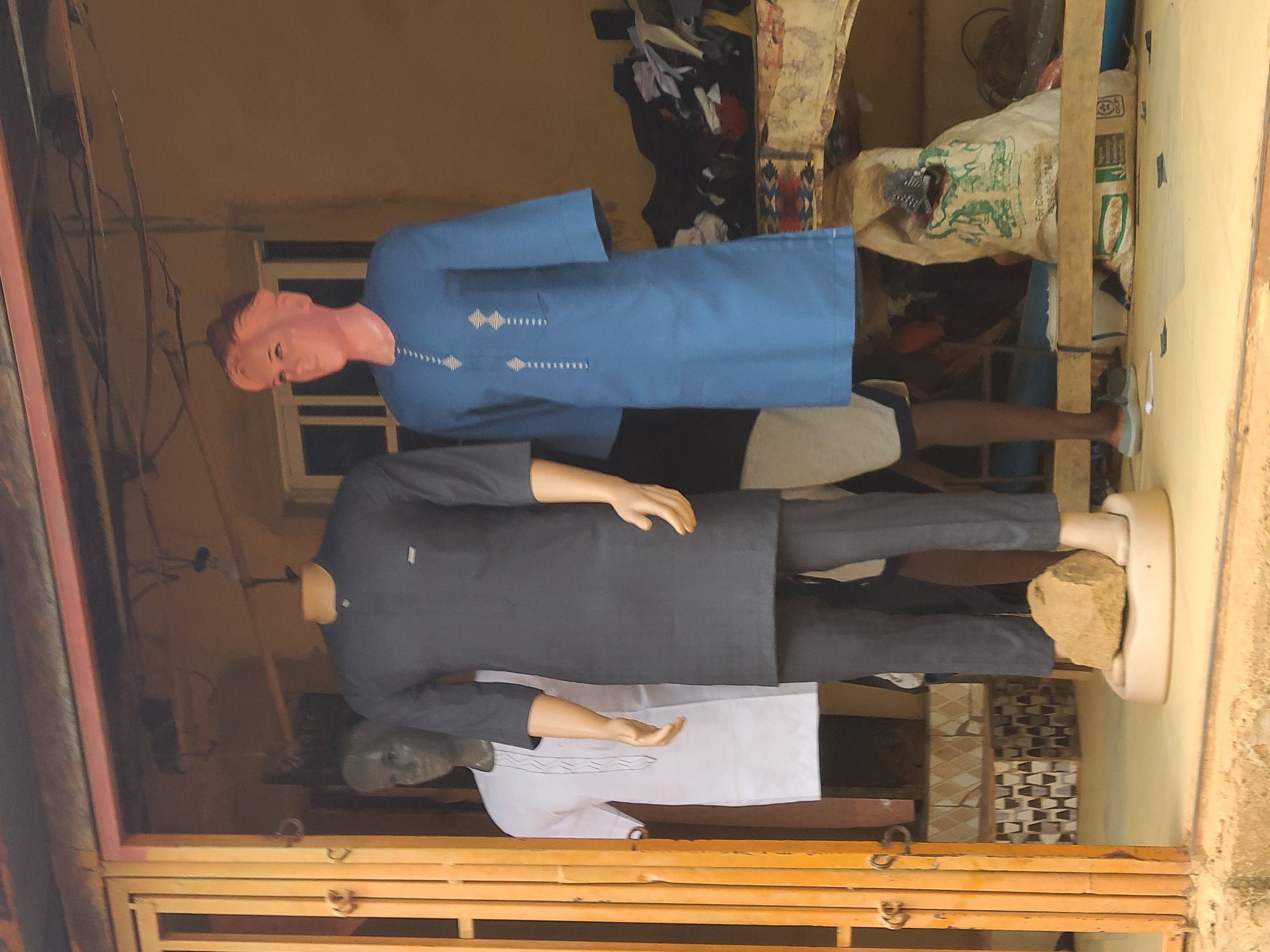 The image shows the entrance to a clothing store or tailor shop in Jos, Nigeria. Three mannequins are displayed in the doorway. The top mannequin is wearing a blue shirt. The middle mannequin is wearing a dark grey suit. The bottom mannequin is wearing a white shirt. The mannequins stand in front of an open storefront with a partially obscured interior. The interior space includes a shelving unit filled with various objects and fabrics, as well as an electric fan. The storefront appears to be located on a busy street. A person stands partially hidden behind the mannequins.