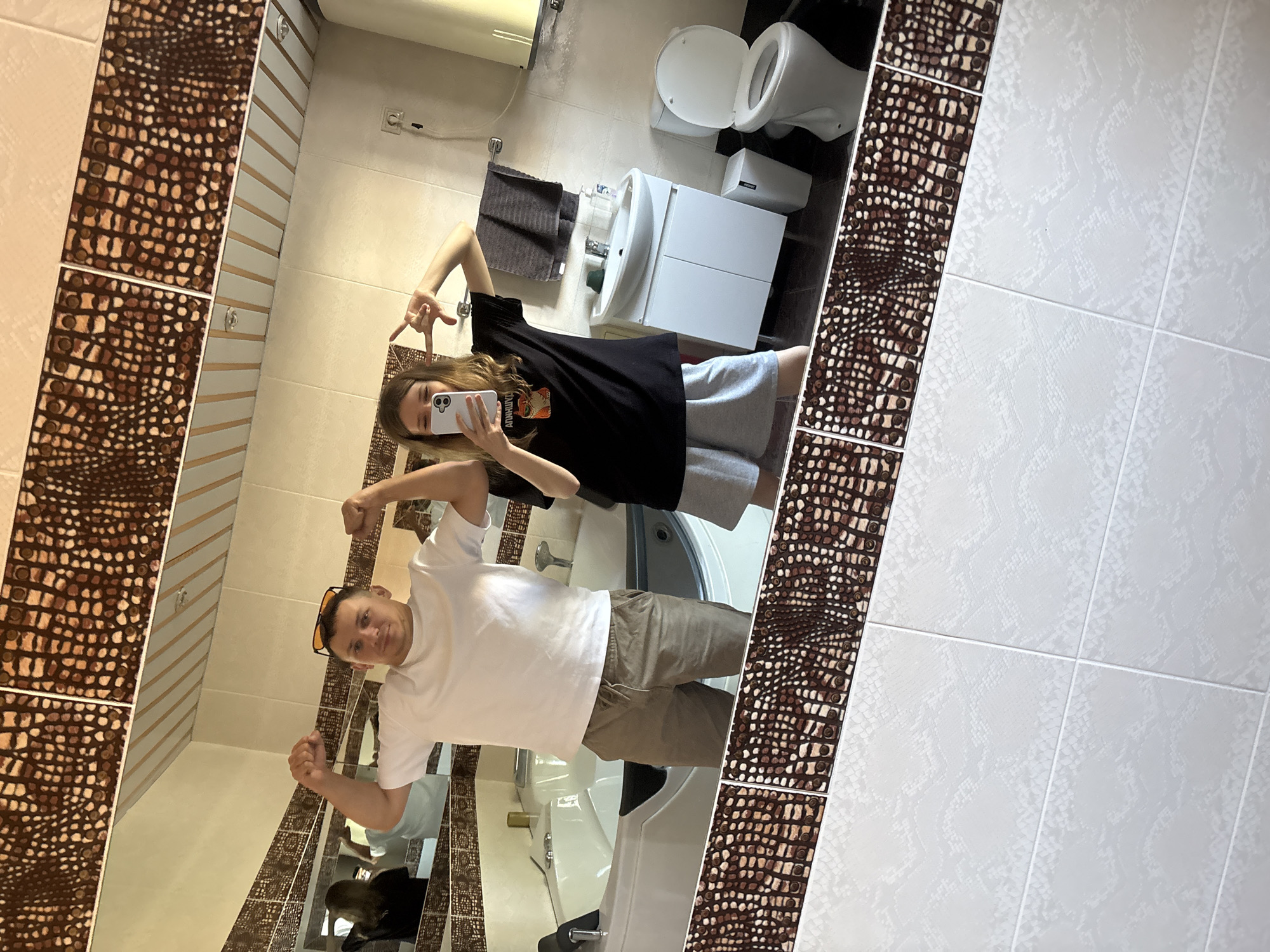 A man and a woman are in a bathroom, taking a mirror selfie. The man, wearing a white t-shirt and khaki shorts, flexes his biceps. The woman, in a black t-shirt and grey shorts, holds her phone up to take the picture and strikes a pose. The bathroom has a large jacuzzi tub, a sink with a white cabinet underneath, and a toilet. The walls are tiled with a brown, textured pattern at the top and a white, scaled pattern on the lower half. The scene is likely indoors, possibly in a residential bathroom in Razdolnoye, Russia, given the context provided, but the image itself does not contain any definitive geographical markers.