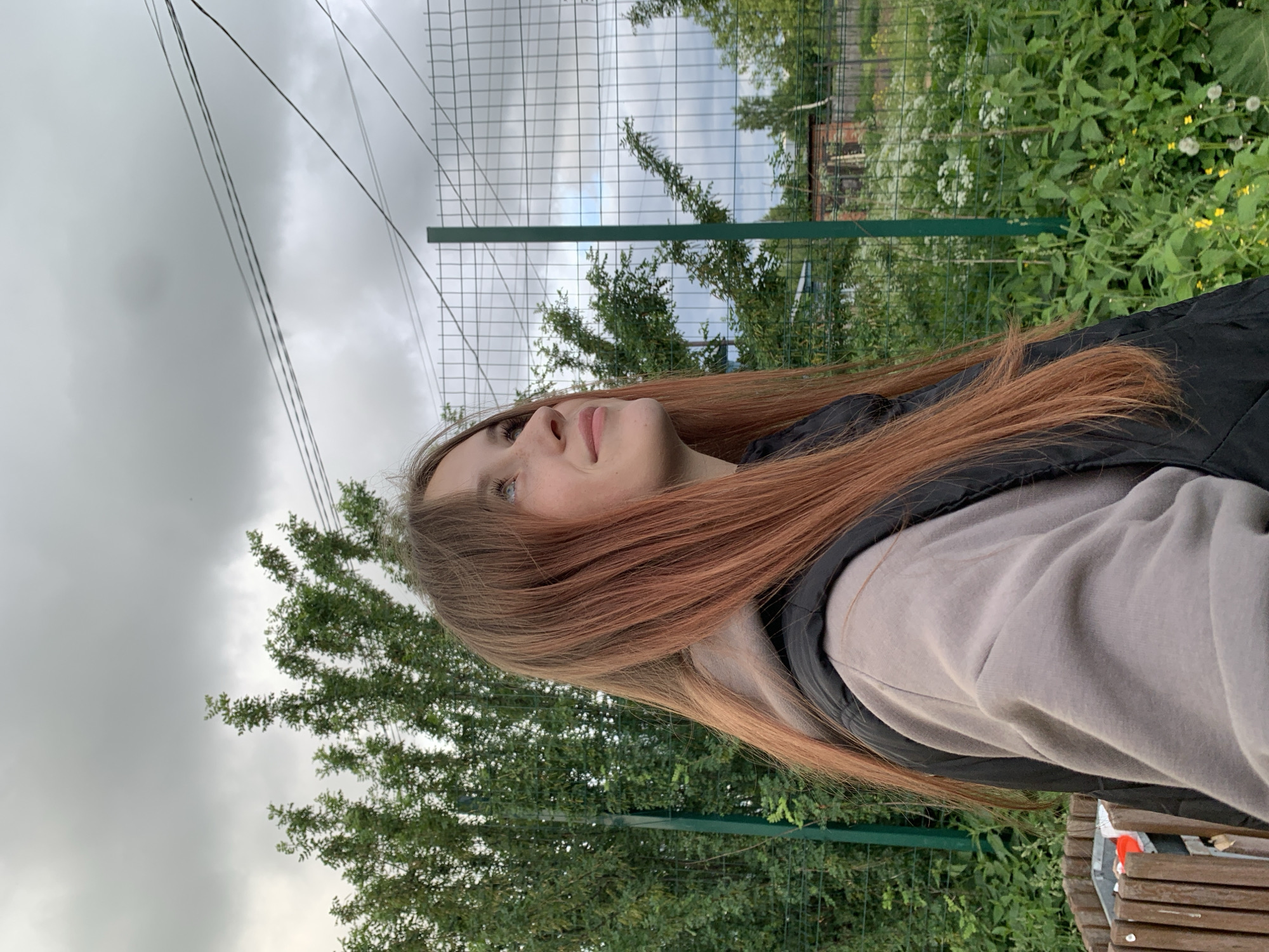 A young woman is depicted in a profile shot, looking upwards and to the right, in an outdoor setting in Фоки, Russia. She has long, straight, reddish-brown hair that falls over her shoulders and down her back. Her face shows a calm, contemplative expression with a subtle, gentle smile, and she has light-colored eyes. She is dressed in a black padded vest worn over a light grey or beige long-sleeved top.
The background is divided by a green metal mesh fence running horizontally across the mid-ground. Below the fence, there is abundant green foliage interspersed with white and yellow wildflowers. Behind the fence, more trees and bushes are visible, and a dark, possibly brick, building can be partially seen through the greenery in the distance to the right. The sky above is overcast and grey, indicating a cloudy daytime. Several black electrical wires or cables stretch across the upper left section of the sky. A portion of a wooden bench or table is visible in the lower right corner of the frame. The woman's pose suggests she is observing something above or in the distance. No visible text is present in the image.
