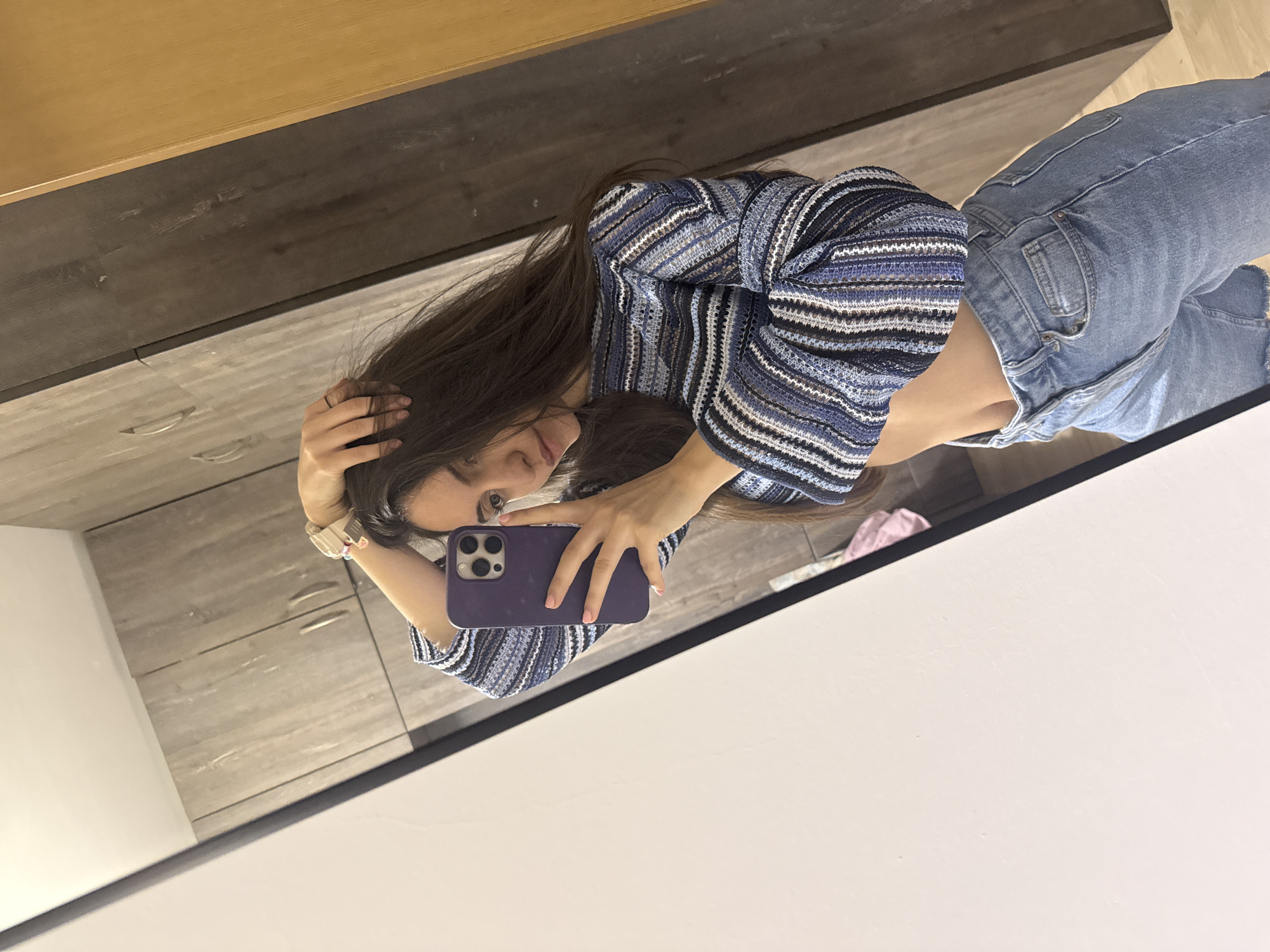 A young woman is taking a selfie in a mirror. She is wearing a striped blue and white top and jeans. Her hair is long and brown. She is holding a purple phone. The background appears to be the interior of a room with a dresser and light-colored walls. The location of the image is unknown. The scene contains a single person taking a selfie with a phone in a domestic setting.