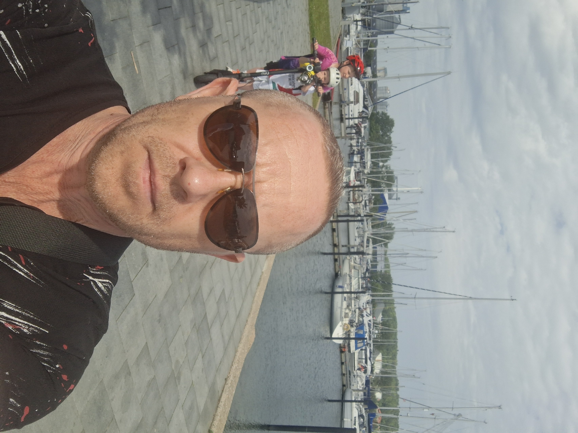 The image is a selfie of a man with sunglasses, taken in Aabenraa, Denmark. Behind him are two children riding scooters. In the background is the harbor with many sailboats. The sky is cloudy. The man is on a paved surface near the water.