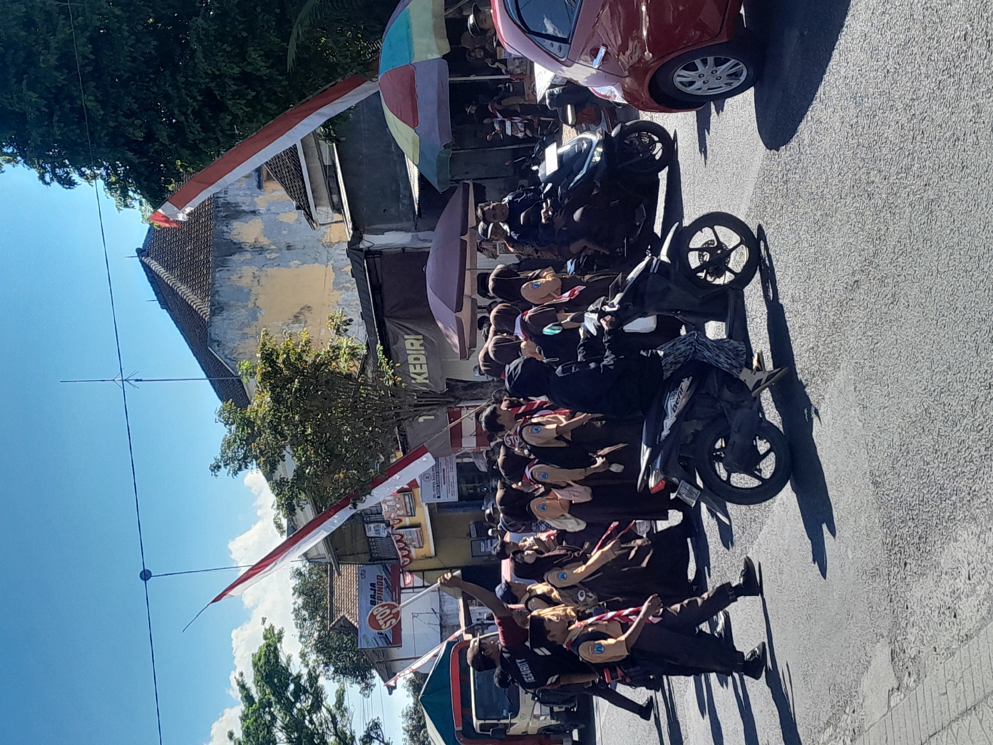 A street scene in Srono, Indonesia, under a clear blue sky features a procession of individuals, many in school uniforms, walking across a paved road. A red and white Indonesian flag is prominently displayed, stretched between poles overhead. Several vehicles, including a white truck, motorcycles, and a red car, are present on the road. Some people are walking, while others are on motorcycles. Buildings line the street, with some showing signs of wear. The overall impression is one of a public event or parade taking place in a moderately busy urban environment.