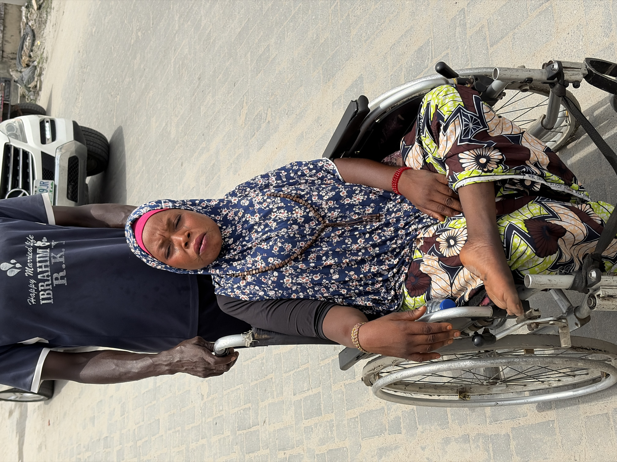 A medium, eye-level shot shows a woman in a wheelchair on a gray brick sidewalk in Igboefon, Nigeria.
The woman has brown skin, a floral headscarf with a pink underlayer, and a floral dress with long black sleeves. She wears a brown beaded necklace and a gold bracelet. Her feet are bare.
A man wearing a navy-blue shirt that says "Happy Married Life IBRAHIM R.K." is pushing the wheelchair. A white vehicle with a license plate that reads "9JR" is parked behind them. The surroundings are blurry and undetailed, suggesting a narrow depth of field.