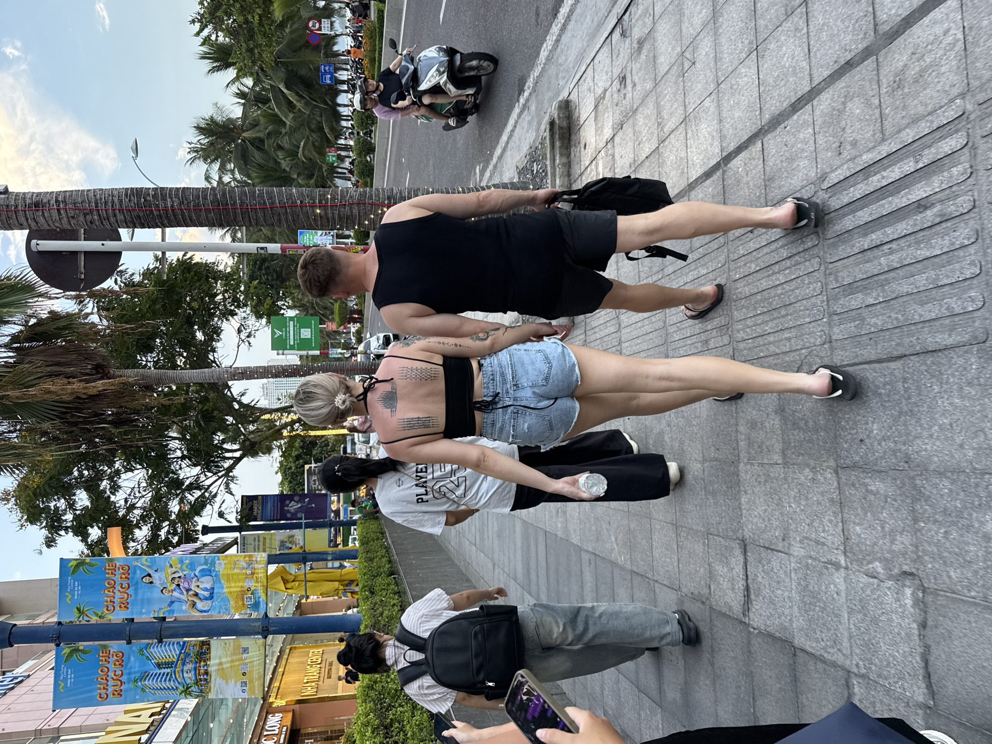 A group of people walk along a sidewalk in Nha Trang Ward, Vietnam. The scene appears to be set in the late afternoon or early evening, with soft, diffused light and a few trees visible. The people are casually dressed, with one man in a black tank top and shorts, and a woman in a black bikini top and denim shorts. Another person in the foreground is wearing a striped shirt and jeans, carrying a backpack. The sidewalk is paved with gray tiles, and there are palm trees and a road with motorcycles in the background. Advertisements for "CHAO HE RUC RO" are visible on banners. The overall mood seems relaxed and unhurried.