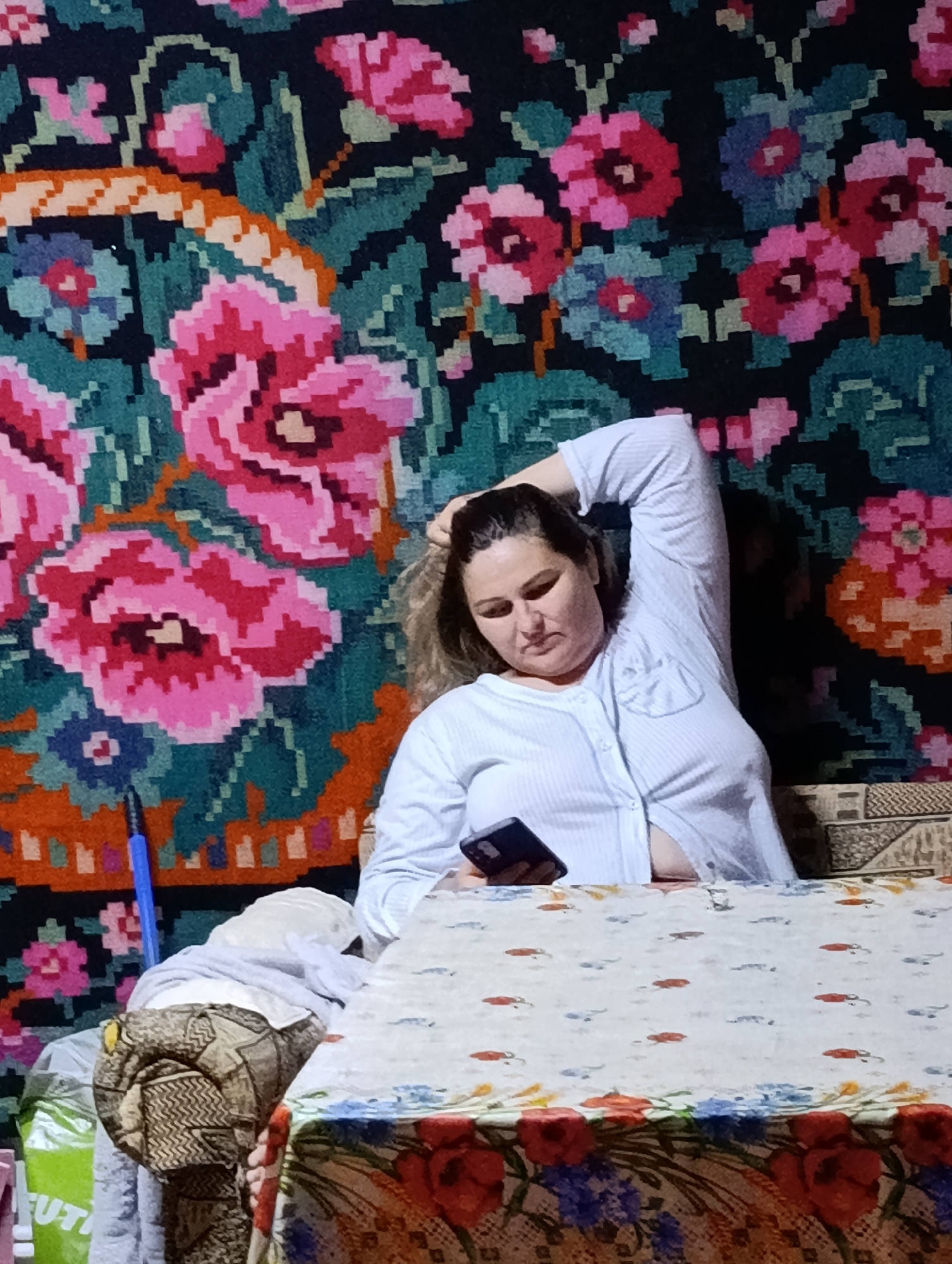 A woman sits at a table in front of a floral tapestry, holding a smartphone. She is wearing a light blue, button-down shirt. Her right arm is bent, with her hand resting on her head. The table is covered with a tablecloth adorned with small red and blue fish. A portion of a couch or chair with a patterned fabric is visible to the woman's left. The tapestry behind her features large, colorful flowers in shades of pink, red, blue, and green against a dark background. The image appears to be taken indoors, possibly in a residential setting in Roghi, Moldova.