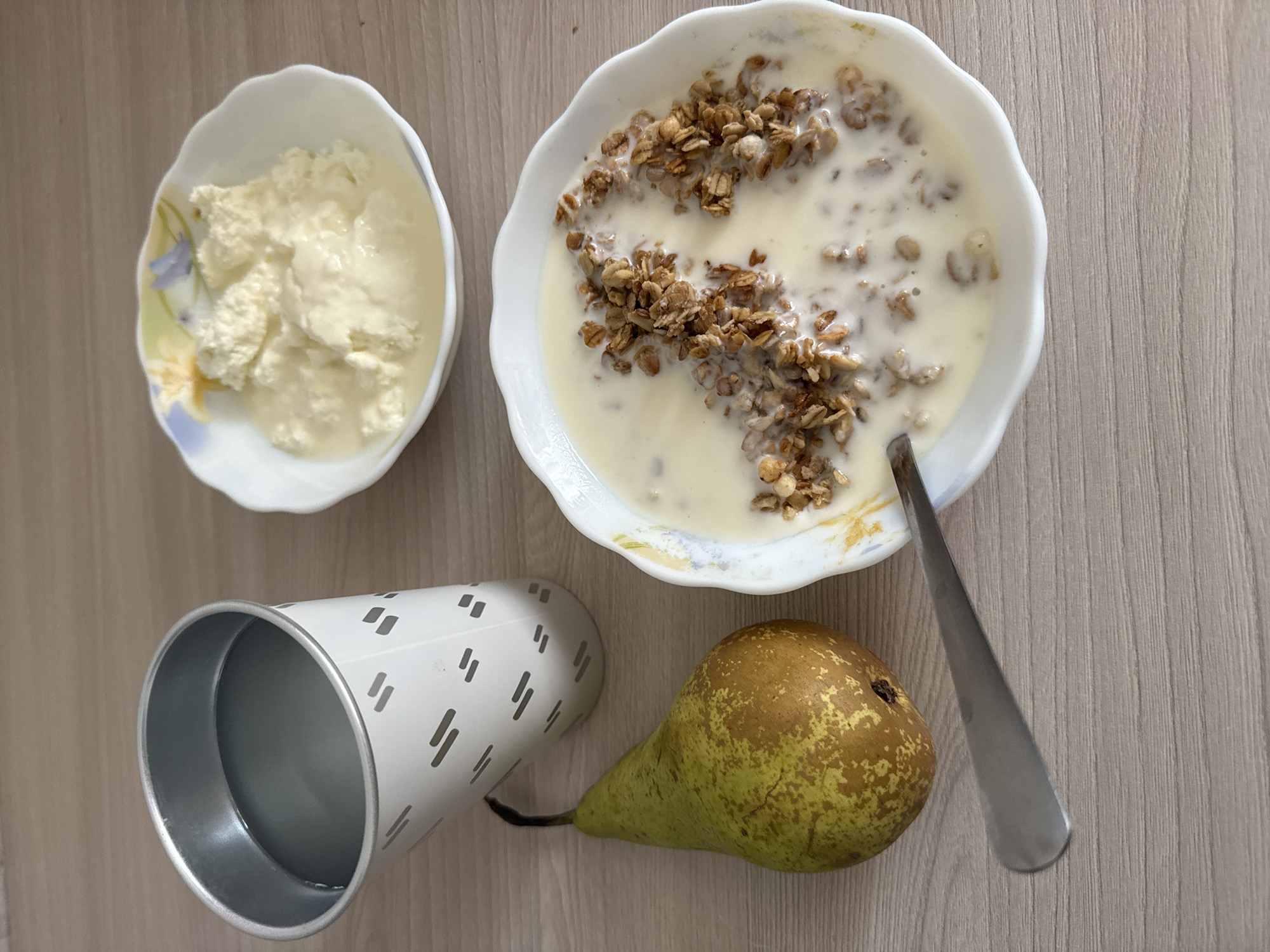 The image shows a close-up, overhead view of a breakfast spread on a light wood-grain surface. On the left, a silver metal cup with a white patterned exterior contains a clear liquid, possibly water or a light beverage. Next to the cup is a green and yellow pear with a brown stem. In the center-right, a white bowl with a floral rim holds a serving of yogurt or cottage cheese with some liquid. Below this bowl, another white bowl, also with a decorative rim, contains a creamy cereal or granola mixture. A silver spoon rests on the edge of the bowl with the cereal. The scene is static and appears to be indoors, with no indication of a specific city or location.