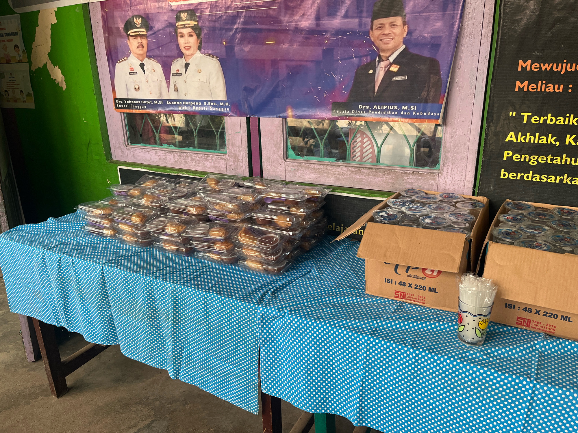 A blue and white polka dot tablecloth covers a wooden table displaying stacks of pre-packaged food items and several boxes of bottled water. Behind the table, a large banner features portraits of two individuals in formal attire, likely government officials, with Indonesian text beneath their images. To the right of the banner, a black board displays text in orange and yellow. The environment appears to be an outdoor setting with a green wall visible on the left. The overall scene suggests a gathering or event where refreshments are being provided in Meliau Hulu, Indonesia.