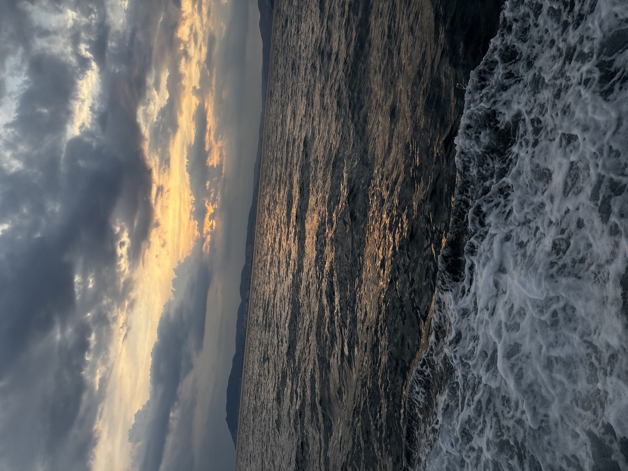 The image depicts a body of water with waves and foam in the foreground, suggesting movement. In the middle ground, the water stretches towards a distant shoreline with hills or mountains. The background features a dramatic sky with dark clouds interspersed with areas of bright, warm light, indicative of a sunset or sunrise. The overall scene is natural and serene, with no human presence or specific urban features visible. The provided information about Kabardinka, Russia, cannot be visually confirmed from the image itself.