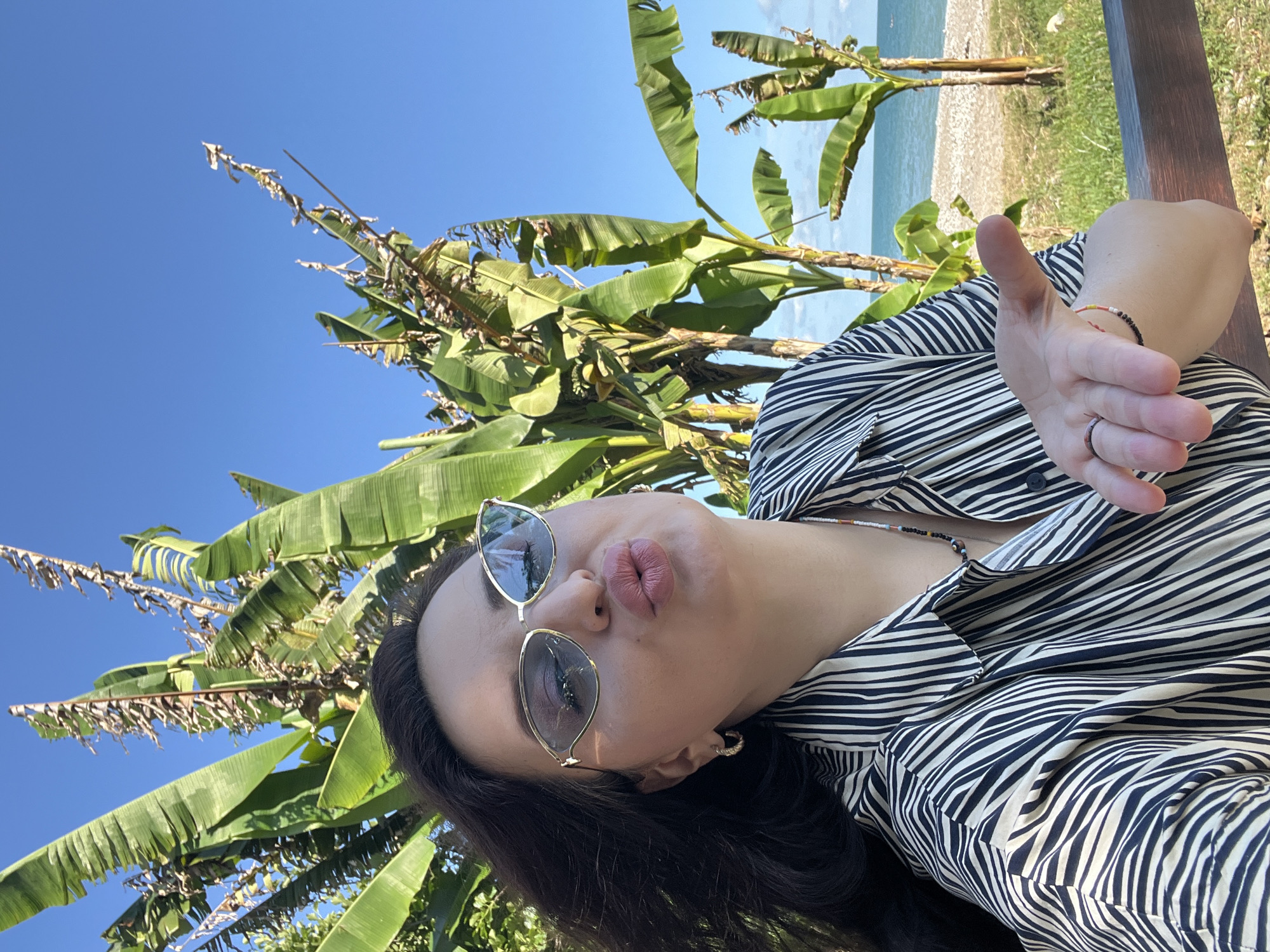 A woman with dark hair is taking a selfie outdoors, leaning forward to offer a kiss. She is wearing sunglasses with gold frames, a black and white striped shirt, and a beaded necklace. She is also wearing earrings and rings on her left hand. Her right hand is extended toward the camera. Behind her, banana trees with large green leaves are visible, and in the distance, a beach and the ocean can be seen under a clear blue sky.