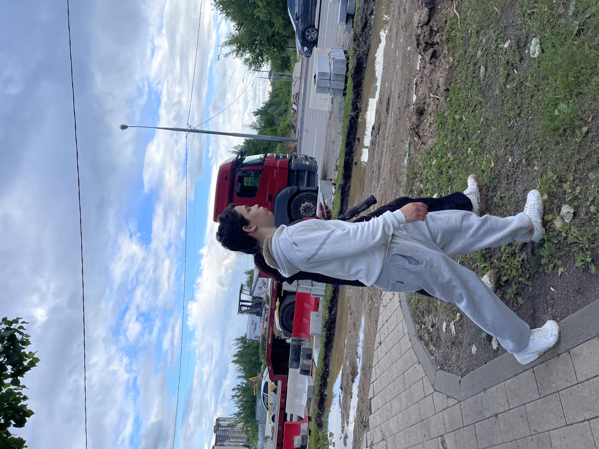 The image shows a street scene in Moscow, Russia, with two young people walking along a sidewalk. The sky is cloudy with patches of blue. In the background, there is a red truck, a taxi, and construction materials, suggesting ongoing road work or development. The ground is wet and muddy in places, indicating recent rain. One person is wearing a white hoodie and light-colored sweatpants, while the other is wearing dark clothing. They are walking away from the camera. The overall scene depicts an urban environment undergoing some form of construction or maintenance.