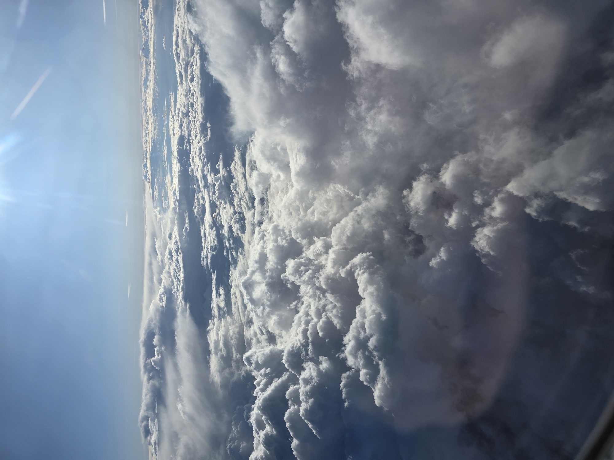 The image shows a view of clouds from above, possibly taken from an airplane. The sky is blue on the left, with the sun visible in the upper left corner. There is a layer of clouds ranging from light, wispy formations to dense, textured formations that are mostly white and gray. It is unknown if this photo was taken above the city of Kommunarka, Russia.