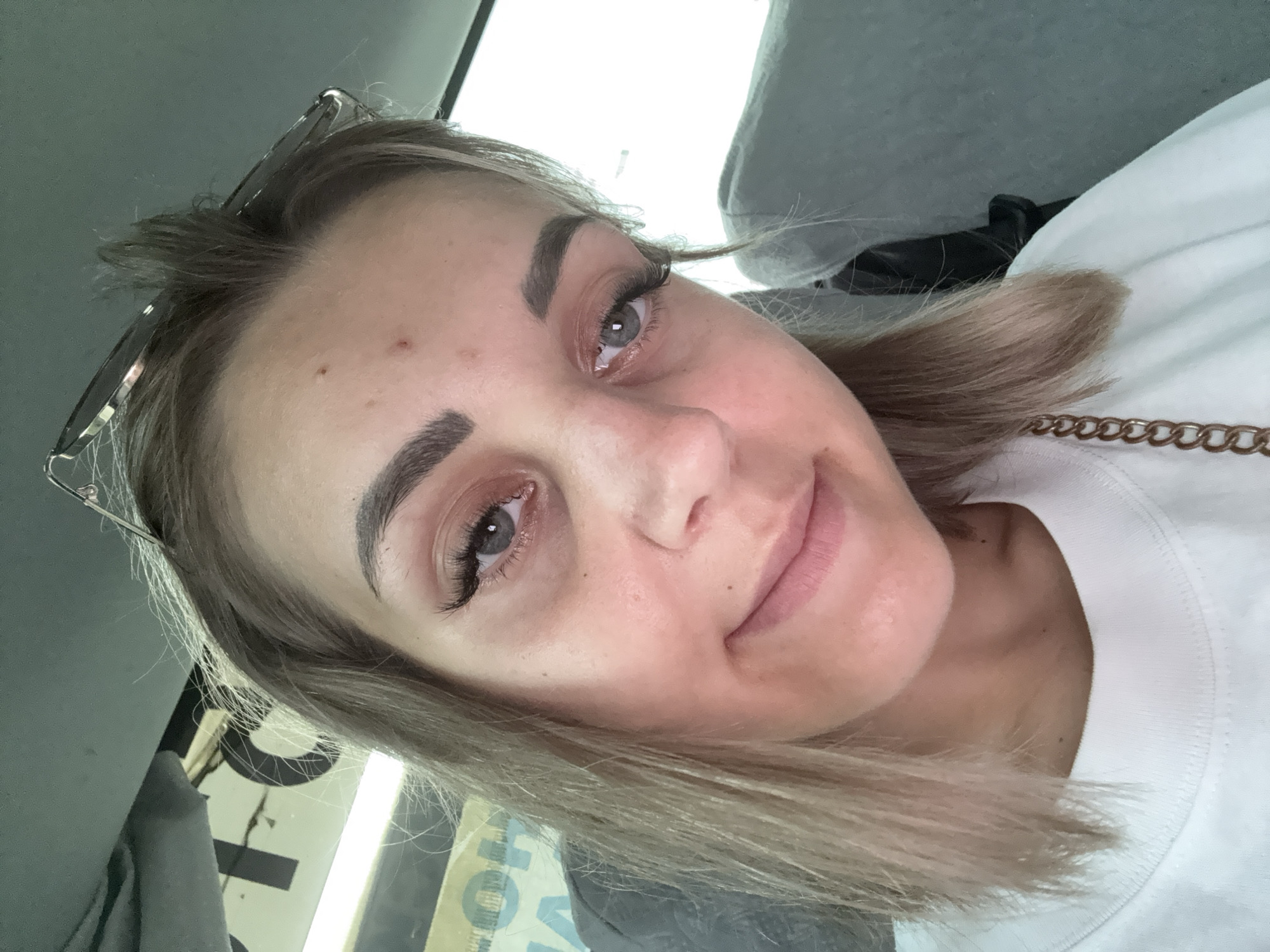 This is a close-up, selfie-style photograph of a young Caucasian woman taken inside a vehicle, likely a bus or taxi, with a muted, natural lighting. She is positioned in the center of the frame, with her head tilted slightly and a subtle smile on her face. Her blonde hair is medium-length and appears to be slightly windswept, with a pair of sunglasses resting on top of her head. She is wearing a white crewneck sweatshirt. Her eyes are blue and appear to have dramatic false eyelashes. There are some minor blemishes on her forehead and under her eyes. The background consists of the interior of the vehicle, including what appears to be a grey seat and a portion of a window. Text is visible on a sign behind her, but it is largely out of focus and illegible. The overall scene suggests a candid moment during travel, with no specific indication of the city of Petropavlovskoe, Russia, beyond the prompt's information.