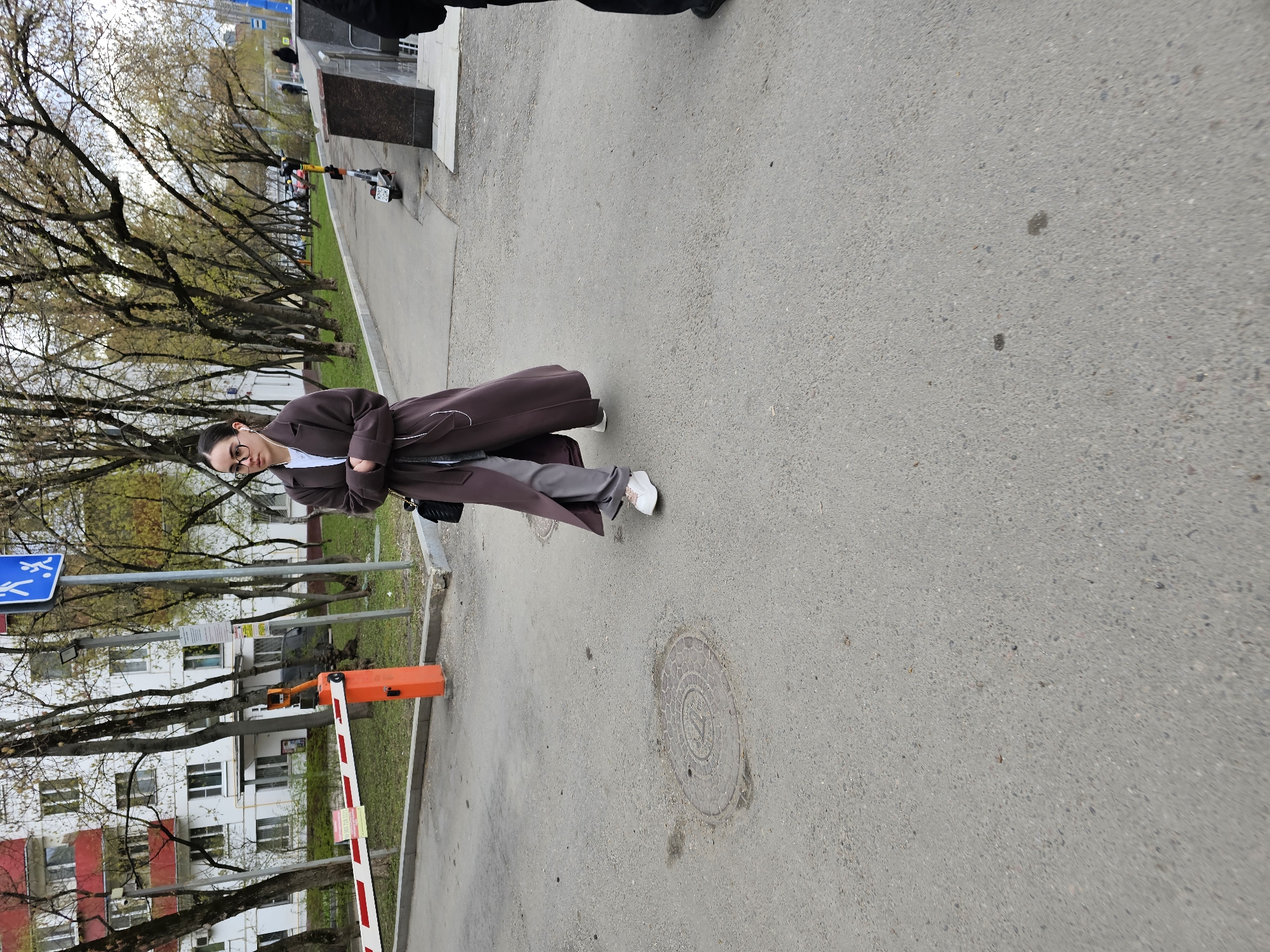 The image displays a young woman standing on an asphalt street in an urban environment, likely in Moscow, Russia, on an overcast spring day. She is positioned slightly to the left of the frame, facing partially away from the camera with a neutral expression, looking slightly downwards.
The woman wears glasses, a white t-shirt, light grey trousers, and a long, dark brown or plum-colored trench coat. She has dark hair tied back and is wearing white sneakers and white earphones. Her arms are crossed in front of her, and a small black bag or wallet is tucked under her left arm.
The background features a line of trees, mostly bare but showing some fresh green buds, indicative of early spring. Behind the trees, multi-story residential buildings, some with white walls and red accents, are visible, characteristic of Soviet-era architecture. To the right of the woman, a grey sidewalk runs alongside the street, and a black electric scooter with yellow accents is parked on it.
In the midground, to the left, a blue sign with white pedestrian figures (suggesting a pedestrian zone) is partially visible. Further left, an orange and white striped boom barrier, used for controlling access to areas like courtyards or parking lots, stands near the buildings. The asphalt ground features two circular manhole covers, one closer to the camera with a hexagonal pattern. The soft, diffused lighting suggests an overcast sky. The only visible activity is the woman posing for the photograph. Text on signs and the scooter is too small or distant to be clearly legible.