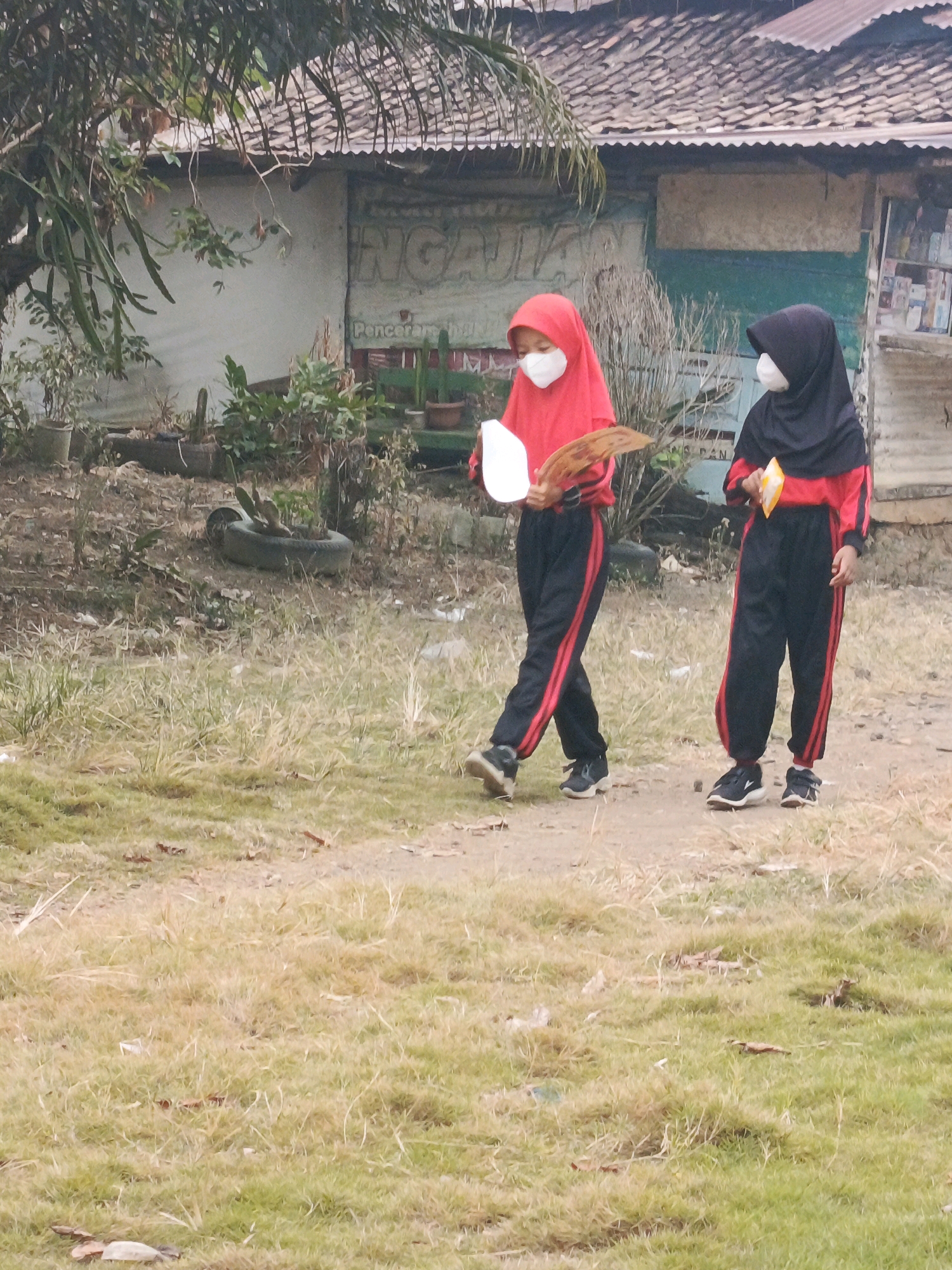 Two young girls, one in a red hijab and the other in a black hijab, wearing matching red and black tracksuits, walk in front of a building. The girl on the left holds a white object and a brown, fan-like item. The girl on the right holds a yellow packet. Both girls are wearing white face masks. The building behind them appears to be a store or small business with various items displayed on shelves. The ground in front of the building is covered in dry grass and dirt. In the background, there are trees and a corrugated metal roof. The scene is located in Megang Sakti II, Indonesia.