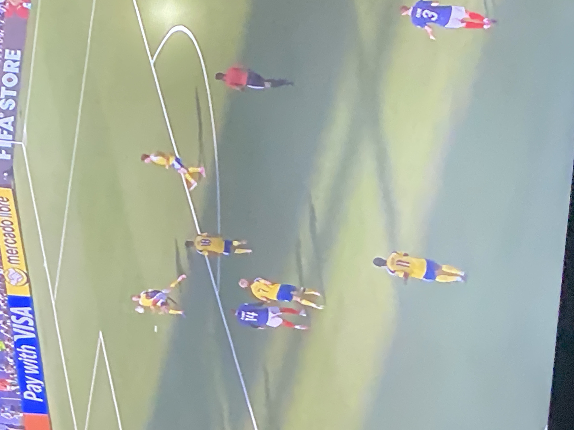 The image shows a soccer match in progress on a grass field with white line markings. Several players in yellow and blue uniforms are visible, as well as players in blue and white uniforms. One player in red is also present. The players are engaged in various actions typical of a soccer game, such as running and kicking the ball. In the background, a banner with the text "Pay with VISA" and "mercadolibre" is visible, suggesting a commercial sponsorship. The scene is outdoors, and the lighting indicates it could be daytime or under artificial lights. There is no information in the image to suggest the location is Zastavna, Ukraine.