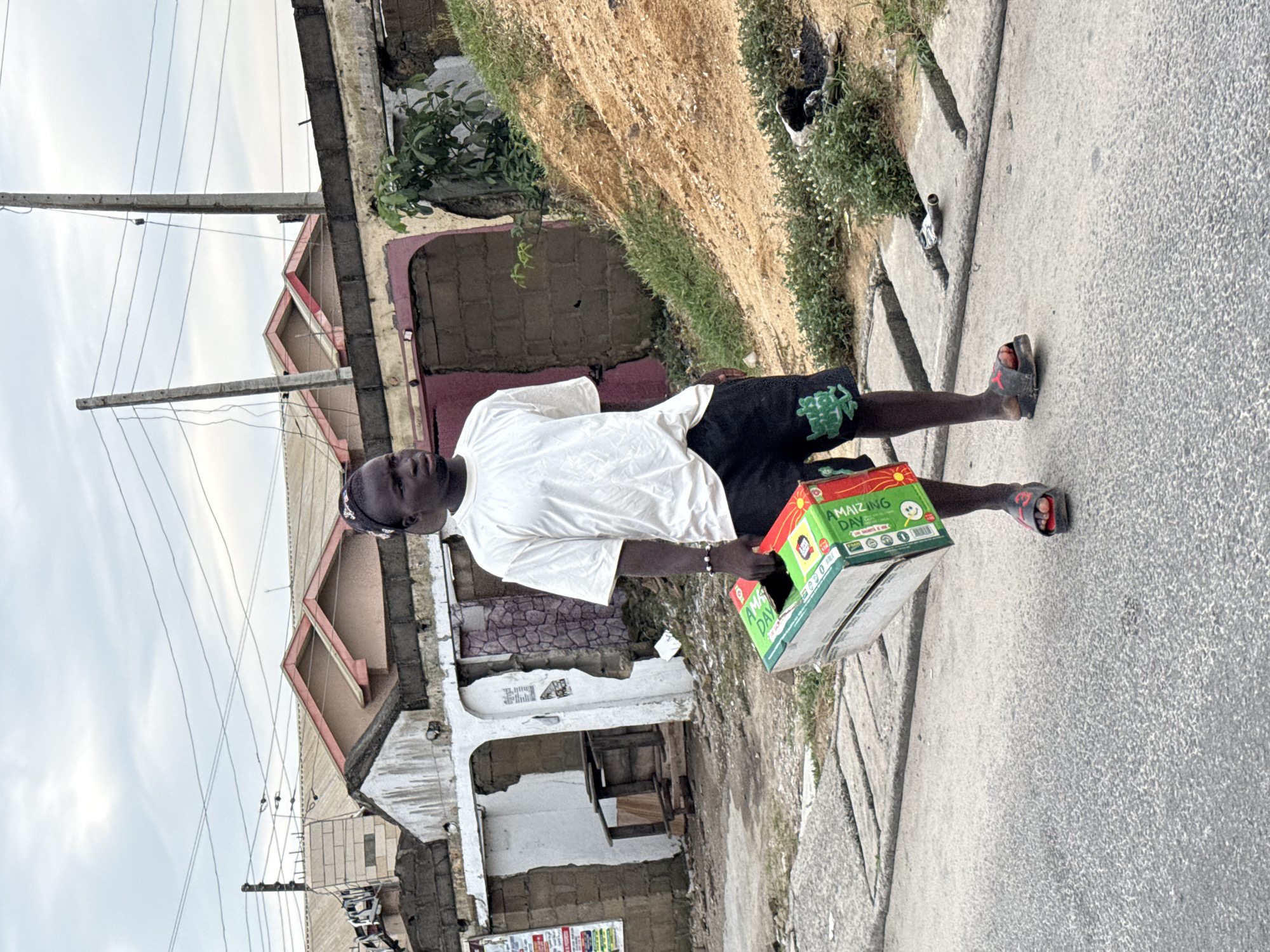A man stands on a sidewalk next to a road in Ado, Nigeria. He's carrying a box that reads "AMAIZING DAY." He is wearing a white t-shirt, black shorts, and sandals. The road is cracked and worn. Buildings with peeling paint and exposed brick line the opposite side of the street. Power lines stretch across the sky. The environment appears urban and slightly dilapidated.