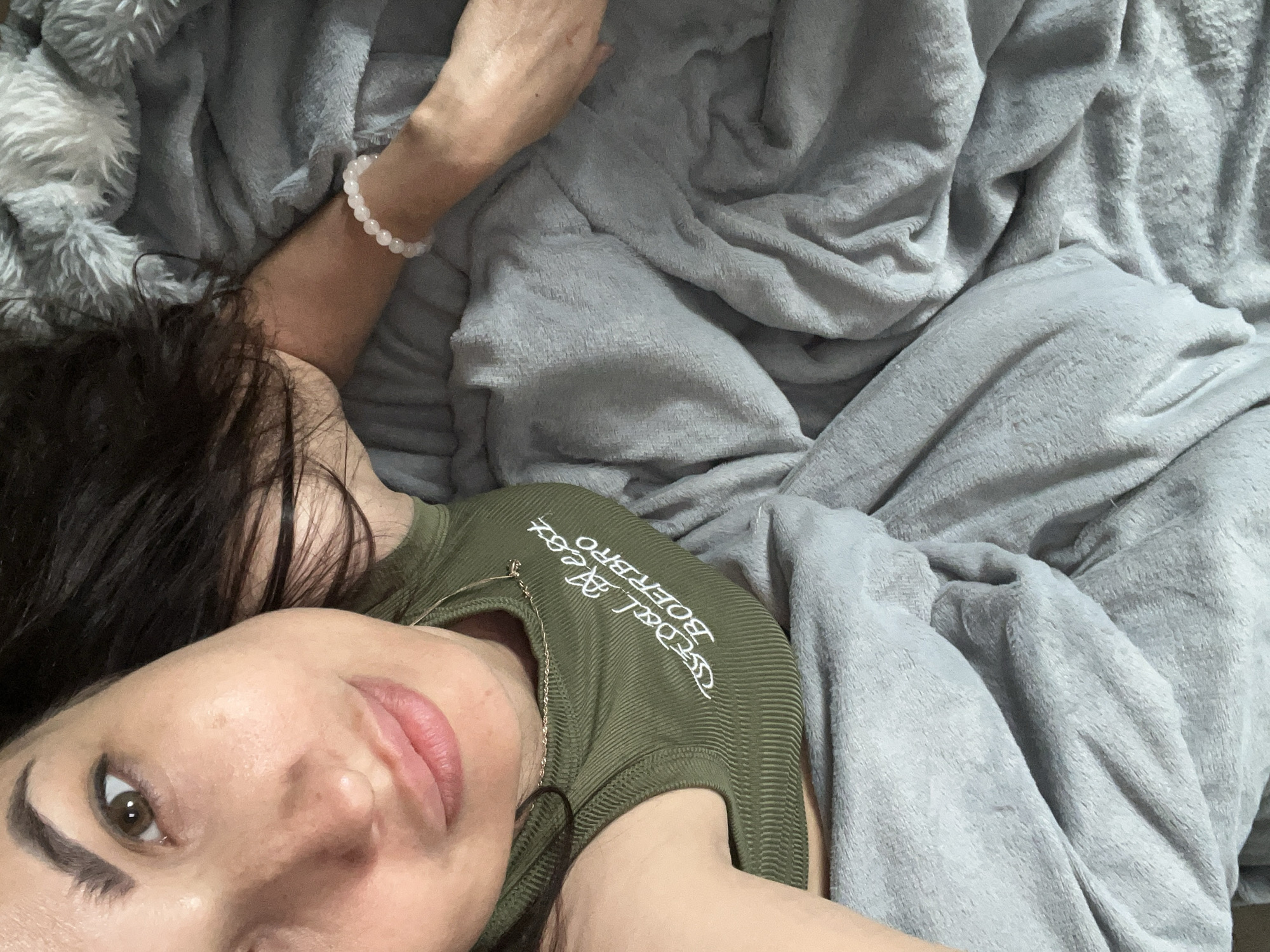 A woman with dark hair is lying in bed, covered by gray blankets. She is wearing a green tank top with white lettering that reads "GOALZ BOERBRO." A delicate gold necklace is visible around her neck, and a bracelet made of small, light pink beads adorns her wrist. The image is a close-up, focusing on her face and upper body. The overall scene suggests a relaxed or intimate moment. The location is unknown.