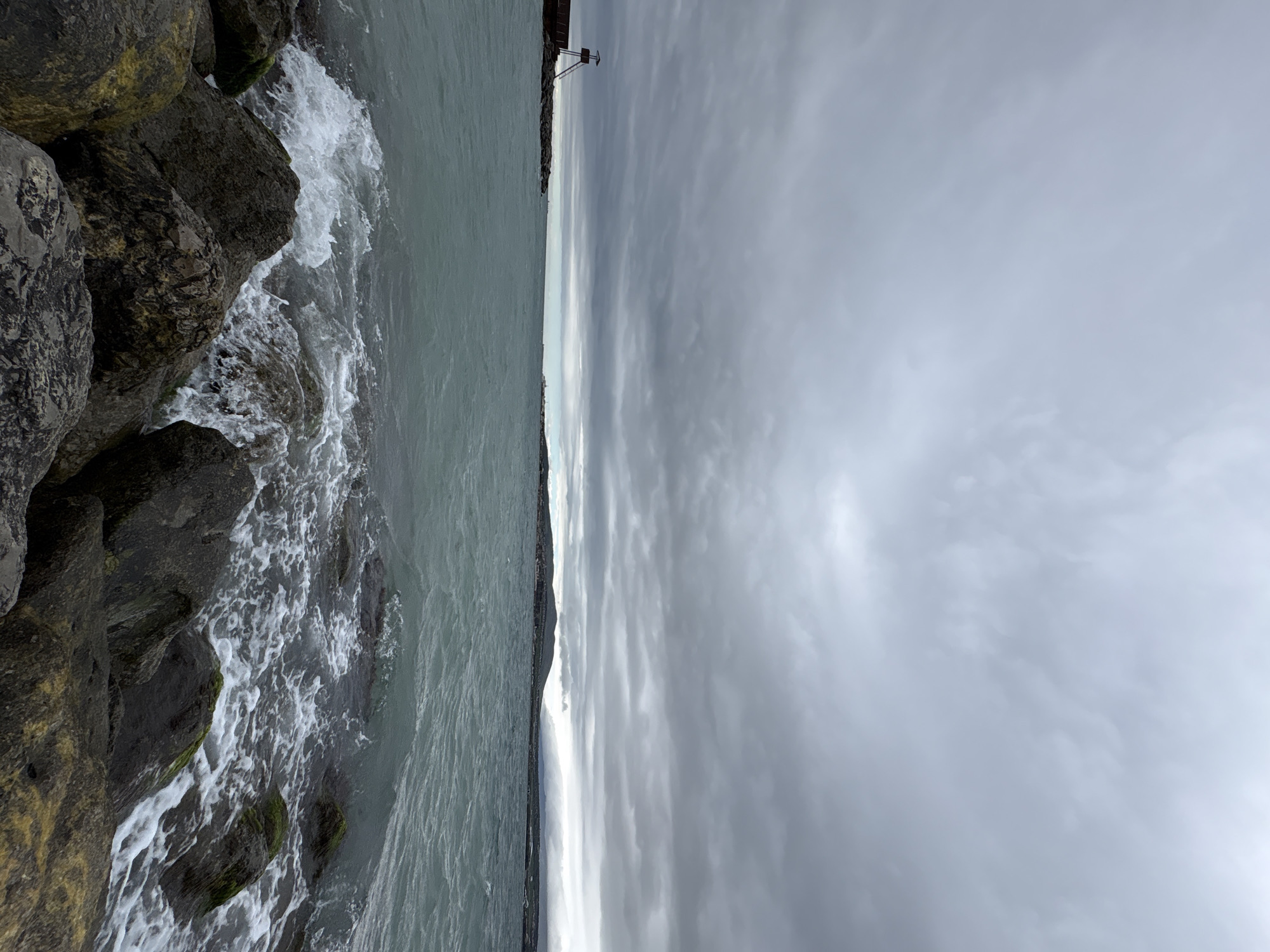 The image shows a coastal scene in Gelendzhik, Russia. On the left, waves crash against a rocky breakwater. The water is a muted green-gray color. A sliver of land appears in the middle ground with what may be buildings, including a tall structure that looks like a lifeguard stand. The right side of the image is dominated by a cloudy, gray sky, providing an overcast atmosphere. The photo seems to be taken at an angle.