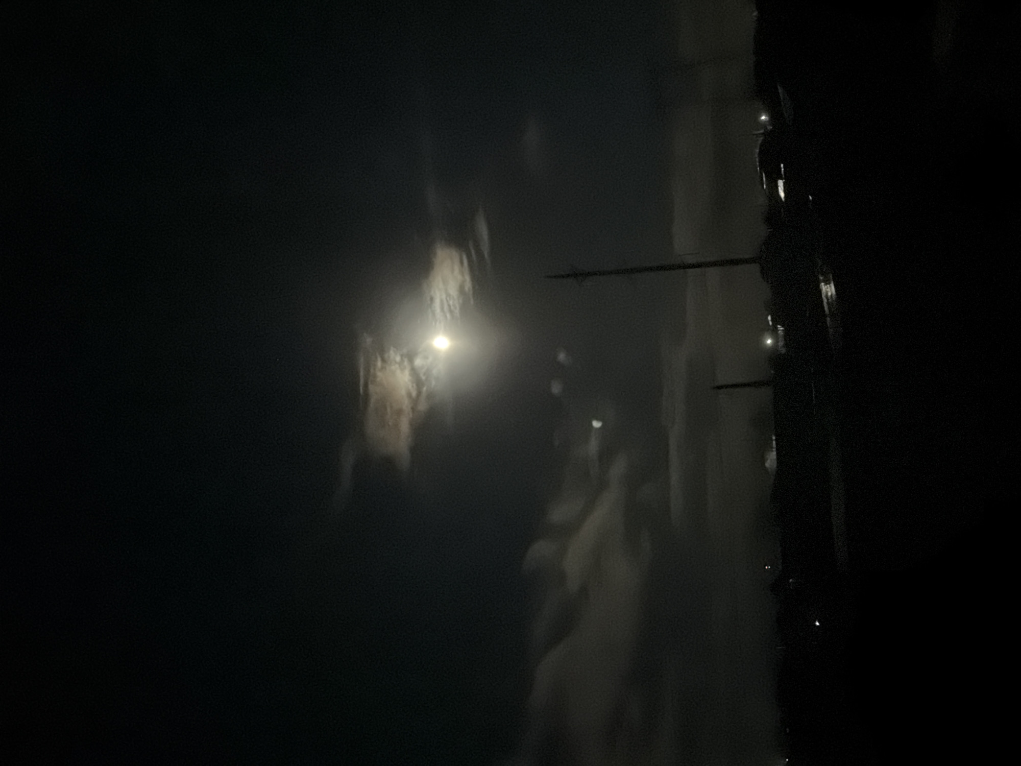 The image appears to be a night shot in Chaikovsky, Russia. The scene is poorly lit, with the primary light source appearing to be a street lamp illuminating a building. The building's facade appears dark. There is a utility pole with wires. The environment is mostly obscured by darkness. No people are visible.